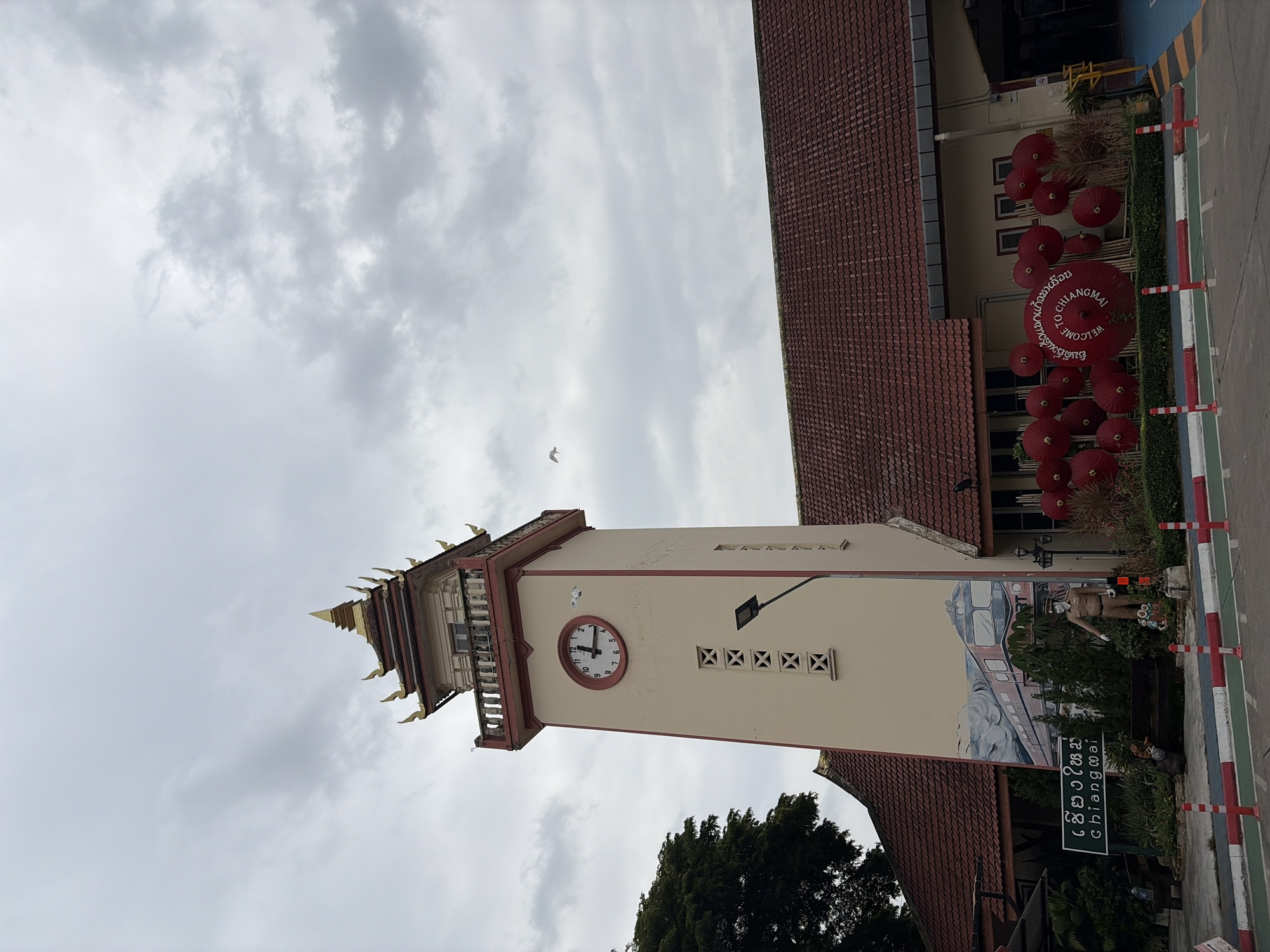 A clock tower stands next to a building with a tiled roof under a cloudy sky. The clock tower is cream-colored with red trim and a gold-colored tiered roof. A round clock face with black hands is visible on one side of the tower. A mural of a train is painted on the lower portion of the tower, and a statue of a person in uniform stands in front of it. A sign in Thai and English reads "Chiang Mai". In front of the building are several red umbrellas arranged in a decorative manner, with some of them displaying the text "Welcome to Chiang Mai". The scene appears to be an outdoor area, possibly a public space or transportation hub. A bird is seen flying in the sky.