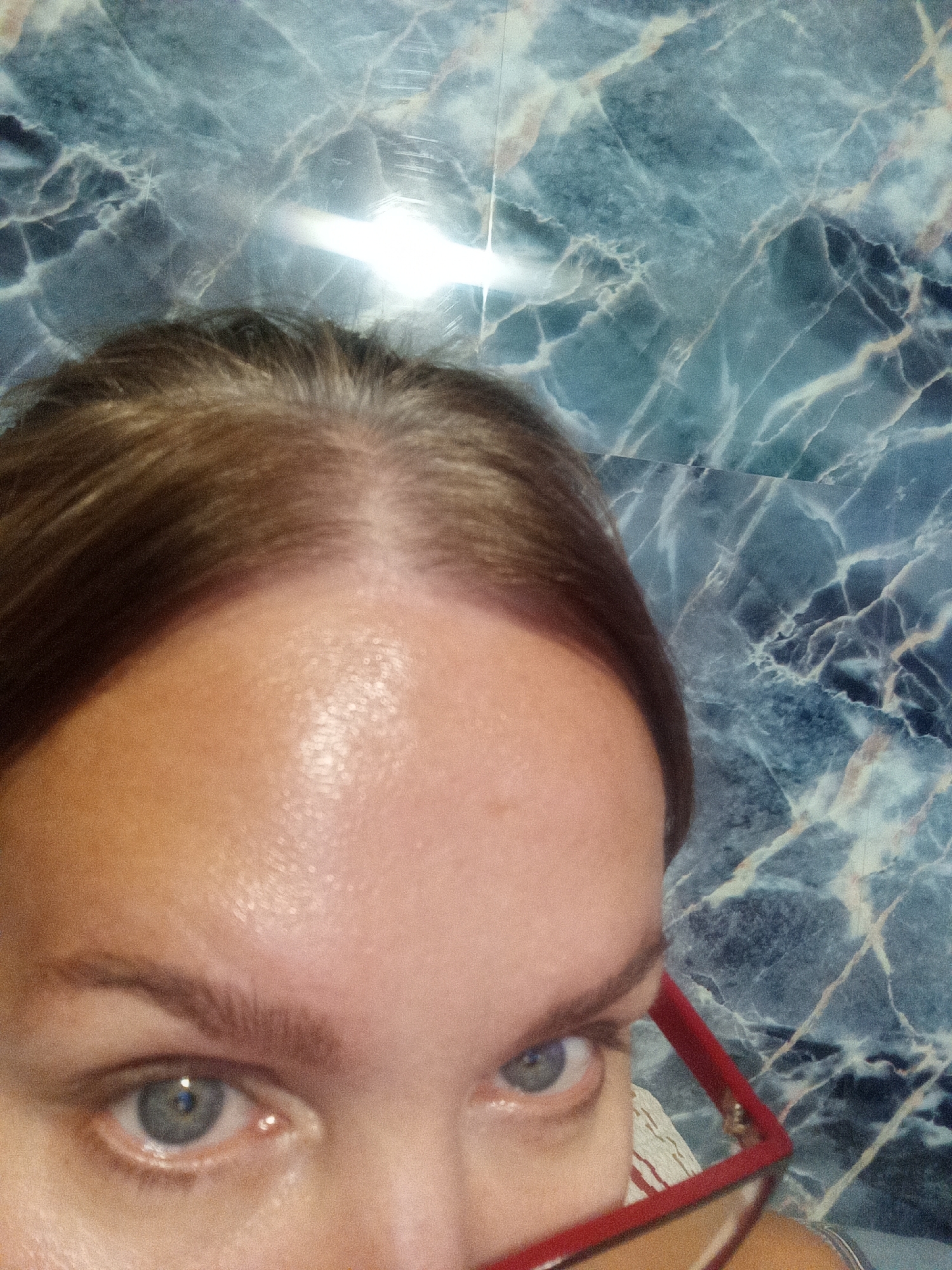 The image shows a close-up of a person's forehead, eyes, and hair. The background is a marbled wall. The person is wearing red-framed glasses. The overall lighting suggests it is indoors, possibly in a bathroom or kitchen given the reflective surface of the wall. There is no identifiable information within the image that would allow for its location to be determined in Novopokrovka, Russia or any other specific location.