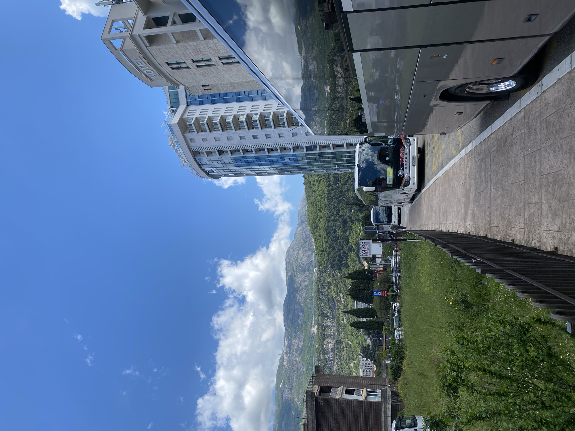 A landscape-oriented photograph captures an outdoor scene, featuring a partially obscured hotel building on the left, and a partially visible large bus on the right. The building is white with reflective blue windows, while the bus is silver.
In the background, a mountain range is visible against a blue sky with scattered clouds. A lush green area with trees and grass separates the foreground and background.
The environment suggests an urban area, possibly a tourist destination, due to the presence of the hotel and bus. The photograph is taken at ground level, and the angle suggests a view from a sidewalk or roadside.
Given the limited information, the location is labeled as Unknown, Unknown.