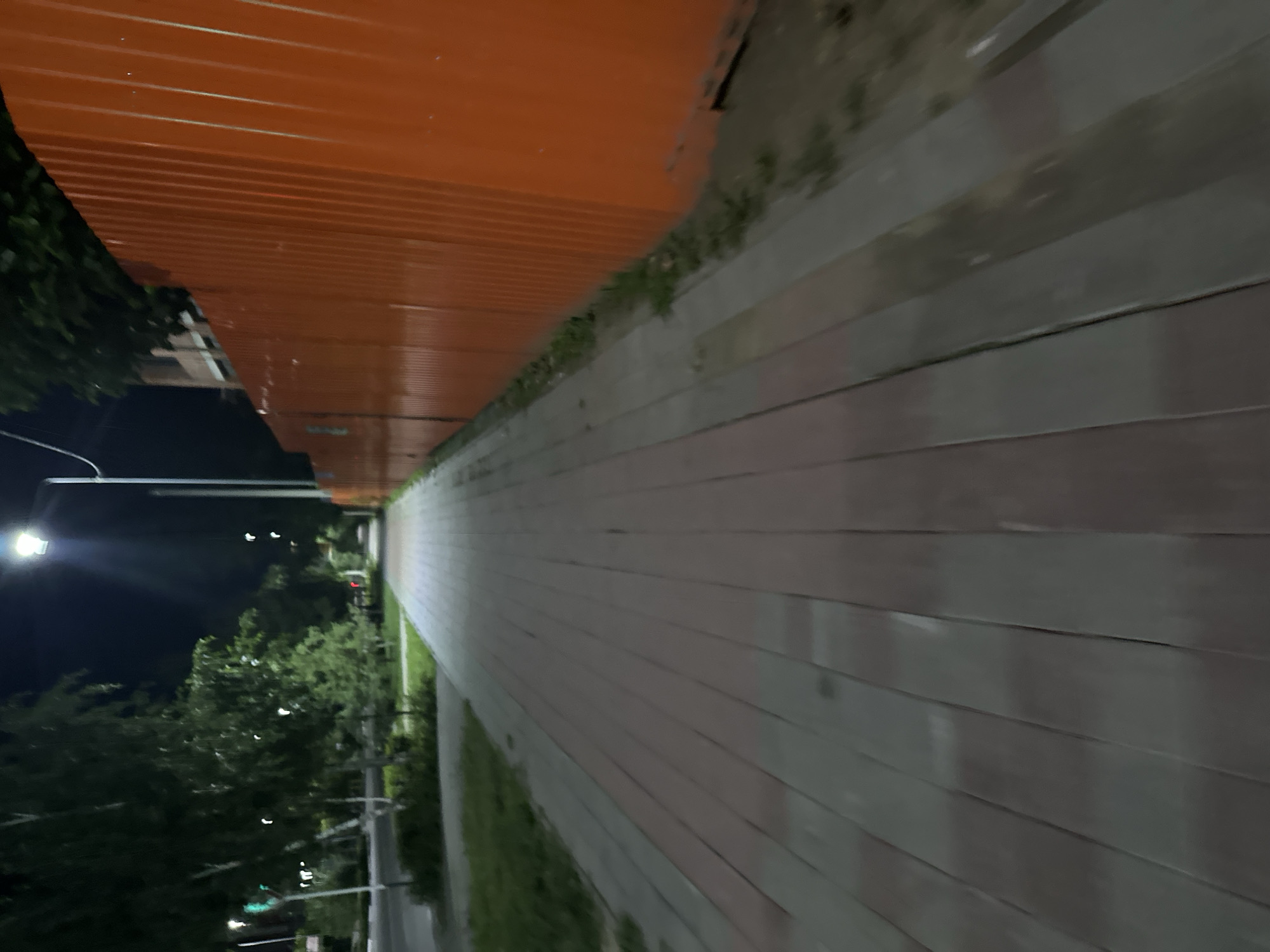 The image depicts a pedestrian walkway at night in the city of Kostanay, Kazakhstan. The walkway is paved with rectangular tiles, alternating between reddish-brown and gray. To the right of the walkway is a tall, bright orange corrugated metal fence, suggesting construction or a perimeter. Beyond the fence, a portion of a brick building is visible, along with trees and the night sky. To the left of the walkway, there is a street with a traffic light showing a green signal, indicating an intersection. Green grass and trees line the left side of the walkway, contributing to a suburban or semi-urban setting. A street lamp illuminates the scene from above, casting shadows and highlighting the textures of the walkway and the fence. There are no people or vehicles visible in the image.