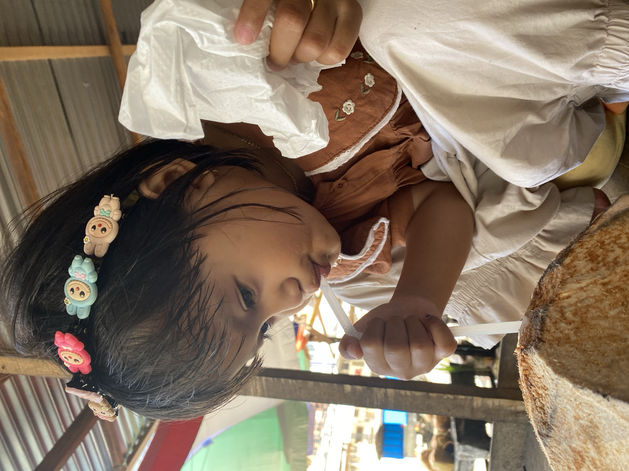 In Ta', Indonesia, a young child, wearing a brown dress with white lace trim and embroidered flowers, and a headband adorned with cartoon animal clips, is seated with a whole coconut in front of them. The child is holding a white straw in their mouth, appearing to drink from the coconut. In their right hand, they hold a crumpled white tissue. The background suggests an outdoor setting with a thatched roof overhead and glimpses of other coconuts and possibly a market stall.