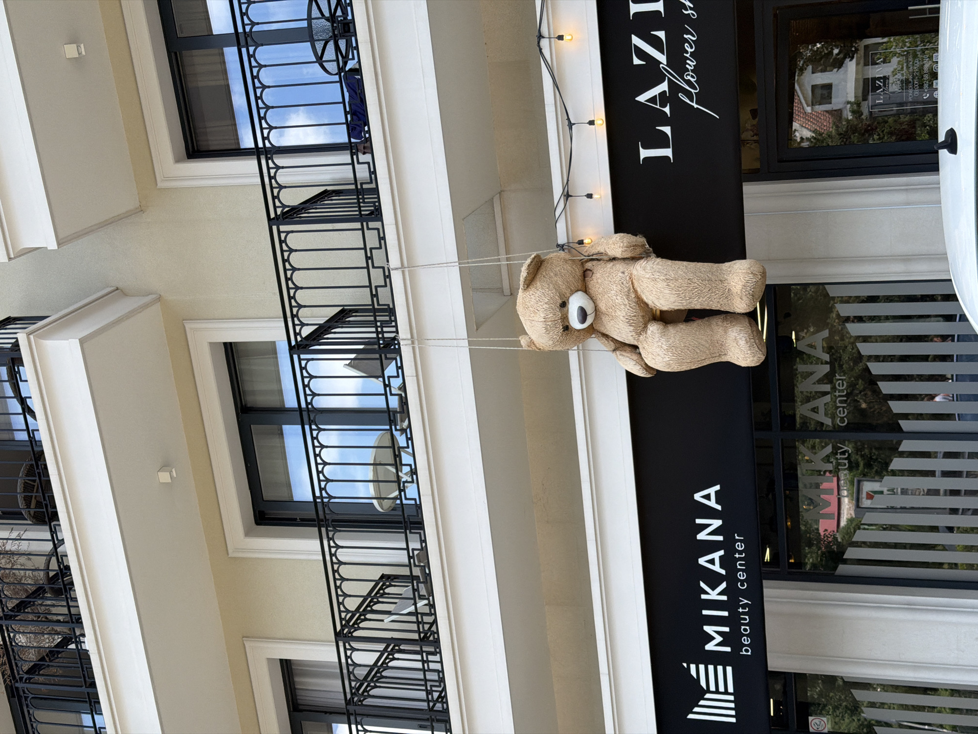 A large, tan teddy bear is suspended from two ropes, appearing to be sitting on a swing, in front of a multi-story building. The bear is positioned in the center of the frame, facing forward with its arms resting on its lap. Above the bear, balconies with black railings are visible on the upper floors of the building. A black awning with white text that reads "MIKANA beauty center" is located below the bear. To the right of the "MIKANA" sign, another awning displays the white text "LAZI flower sh". The building appears to be a commercial establishment, likely in an urban setting. The scene is brightly lit, suggesting daytime. The lower portion of the image shows the reflection of trees and a red-roofed building in the glass storefronts. A white car is partially visible in the foreground. The location is identified as Budva, Montenegro.