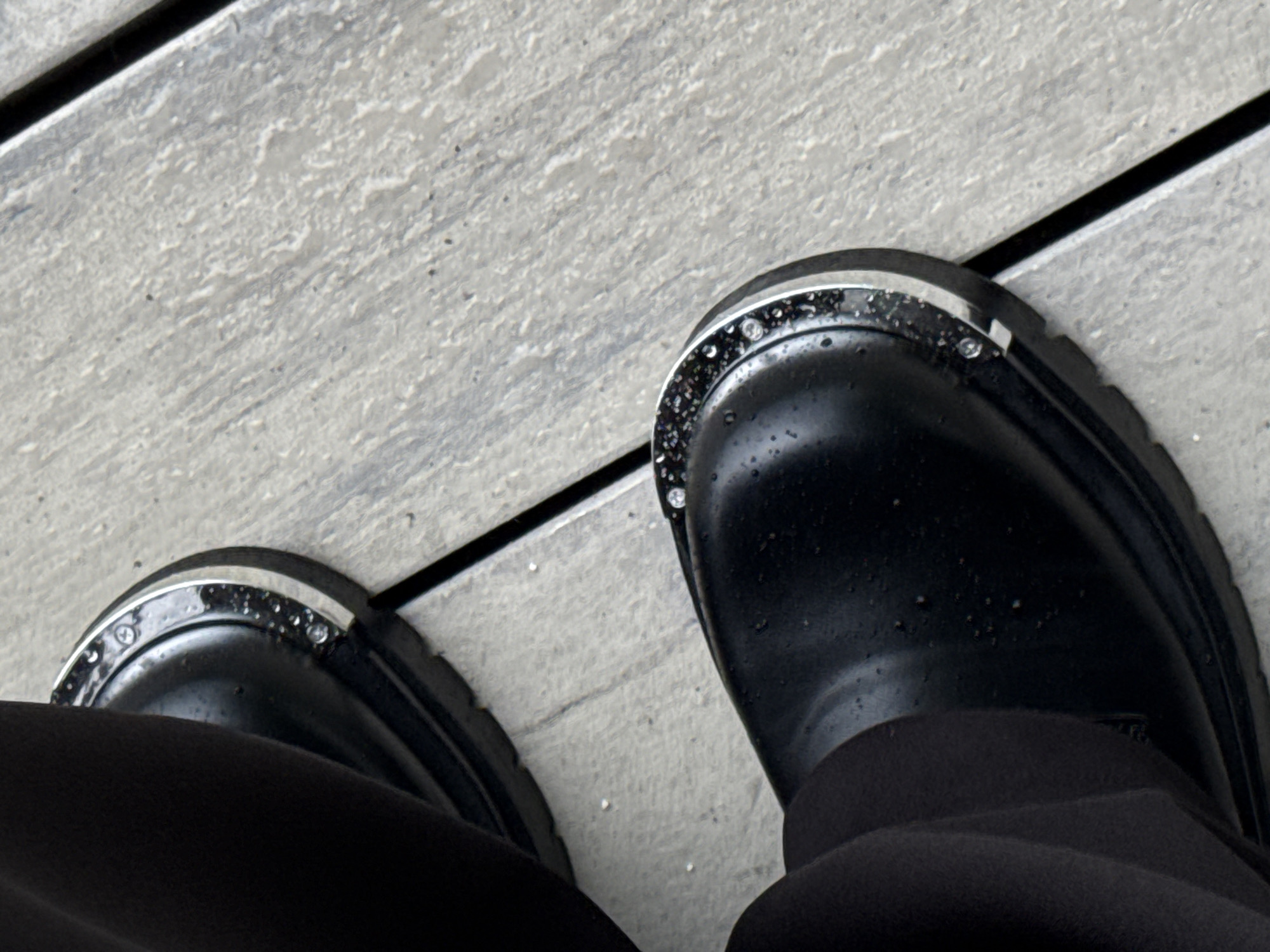 The image is a high-angle, close-up shot of two black, chunky boots standing on a light grey, textured surface that appears to be made of wood planks or concrete with a wood grain pattern. The boots have a metallic trim around the toe area, with visible screws or rivets securing it. Small droplets of water are scattered across the surface of the boots, suggesting recent rain or damp conditions. The setting is outdoors, given the textured ground and the presence of water droplets. The lighting suggests it could be daytime, possibly overcast or late afternoon due to the lack of strong shadows. The image is taken from a perspective looking down at the feet, implying the subject is standing or has just finished walking. No text is visible in the image. The overall mood is neutral, focusing on the footwear and the immediate environment. The location context provided is Votkinsk, Russia, which could imply a cooler climate where boots are practical footwear.