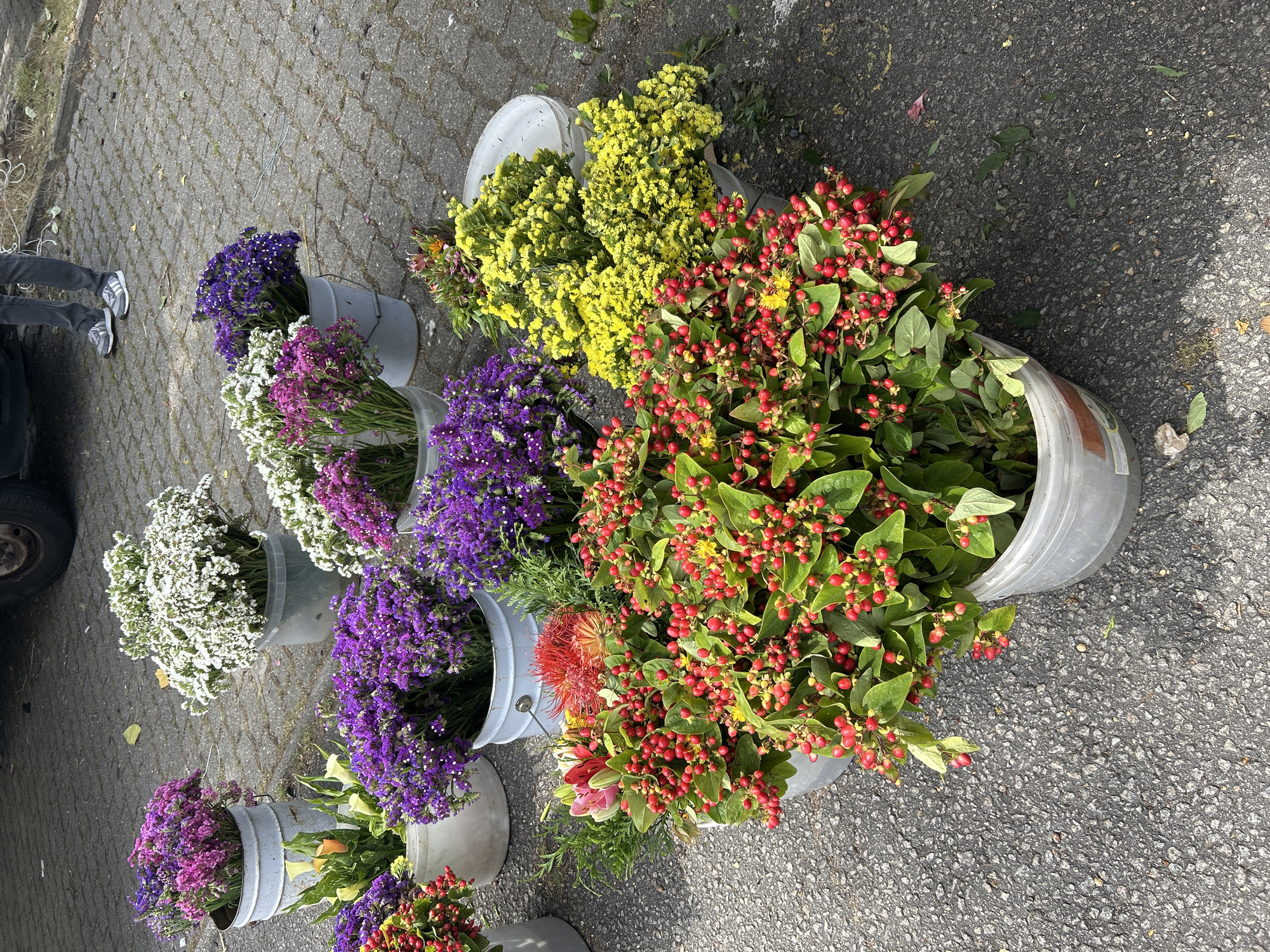 A market stall displays buckets of various flowers on a paved surface. The flowers include bunches of vibrant red berries with green leaves, yellow clusters, purple blossoms, and white and pink blooms. In the background, the legs of a person wearing gray jeans and athletic shoes are visible, standing near a vehicle. The scene appears to be outdoors in a public space.