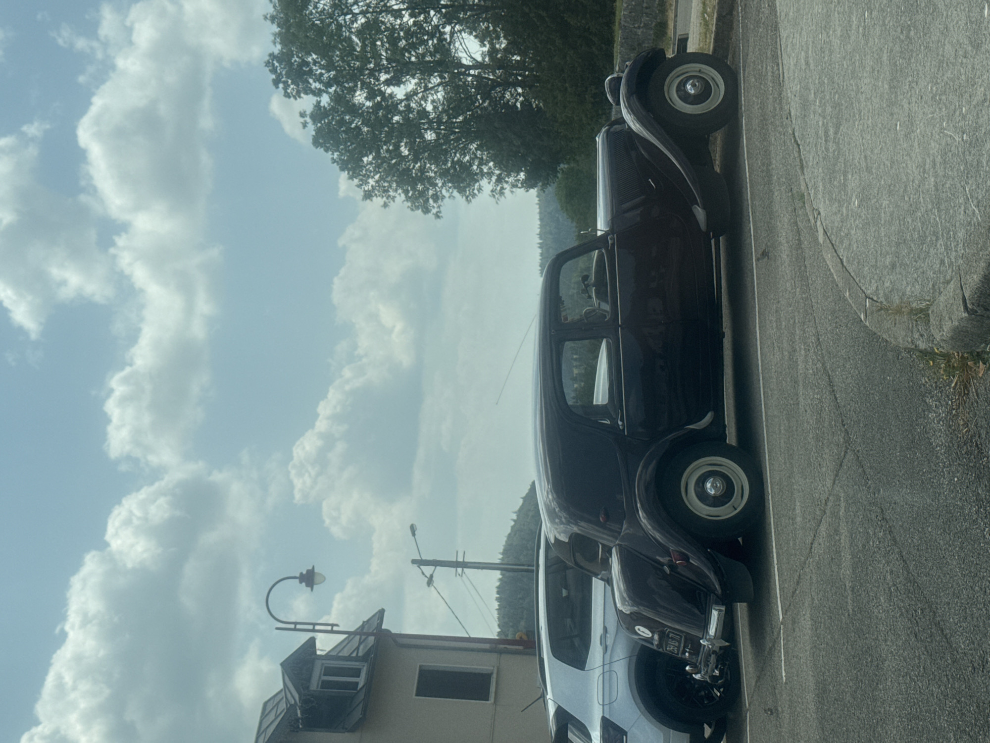 In La Chaux-du-Dombief, France, a dark maroon vintage Citroën car is parked next to a modern white SUV in a paved parking lot. The Citroën, with its distinctive curved fenders and elegant silhouette, is positioned in the foreground. The white SUV is slightly behind and to the left of the vintage car. In the background, a building with a peaked roof and a window is visible to the left, and a streetlamp stands nearby. Further back, a line of trees and a hillside create a natural backdrop under a sky filled with white, fluffy clouds. The scene appears to be a still, outdoor environment during daylight.