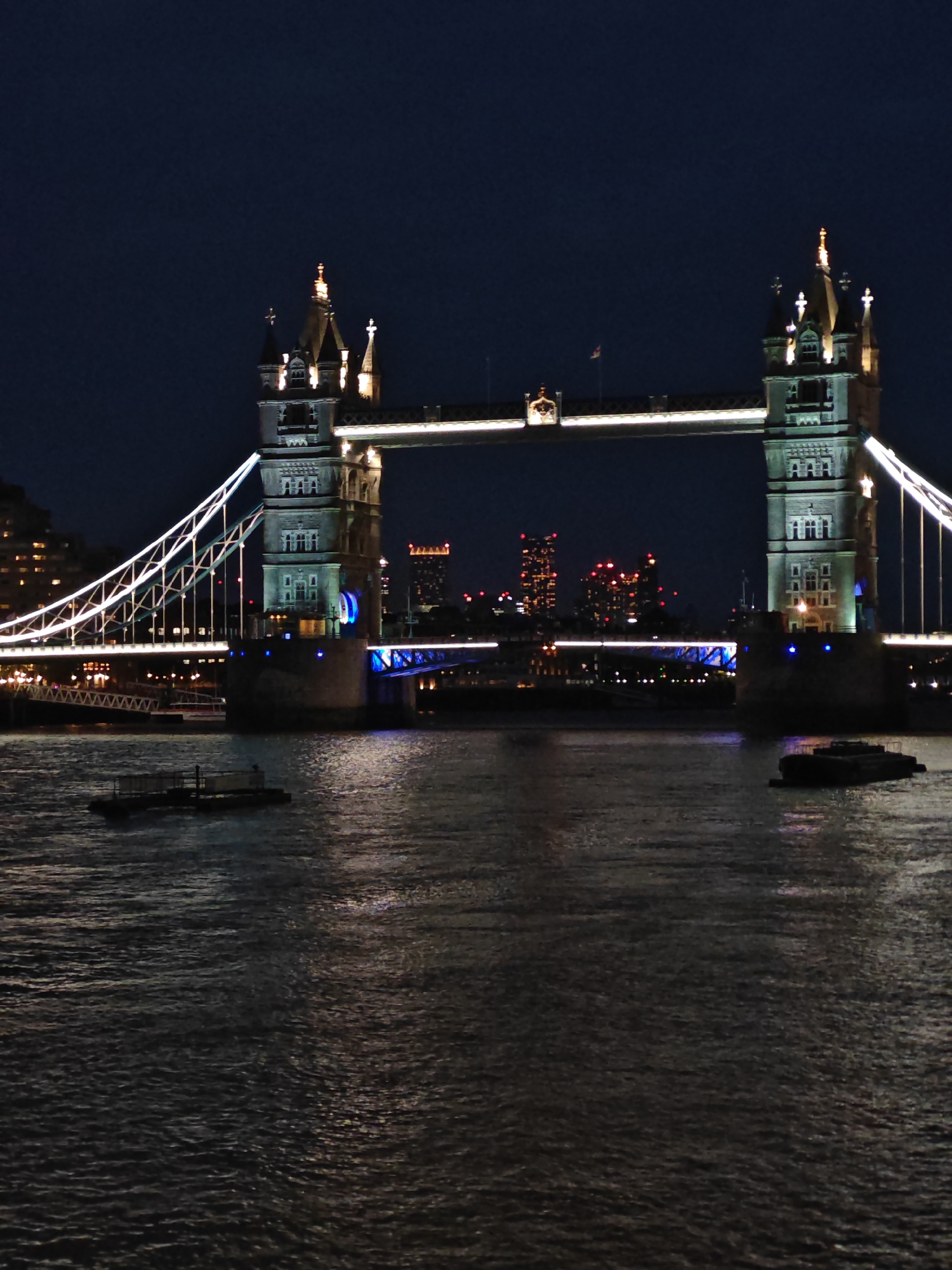 This image depicts the Tower Bridge in London, United Kingdom, at night. The bridge is illuminated with white and blue lights. The Thames River is in the foreground, reflecting the lights of the bridge and nearby buildings. Two barges are visible on the river. In the background, city buildings are dimly lit.