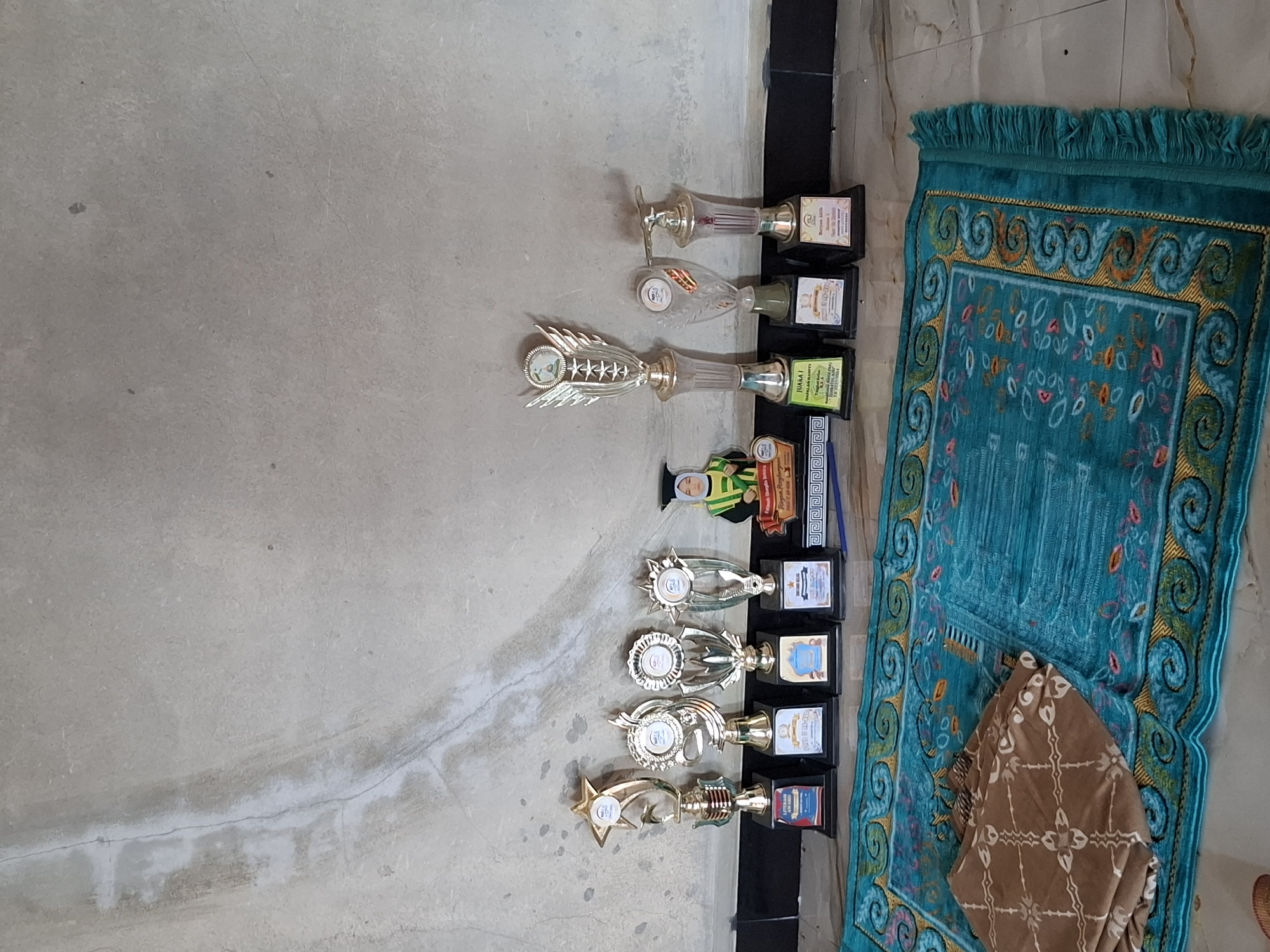 A framed diploma is displayed on a rug in front of several trophies. A wall with a concrete texture serves as the backdrop. The image is a street view in Gronggrong, Indonesia.