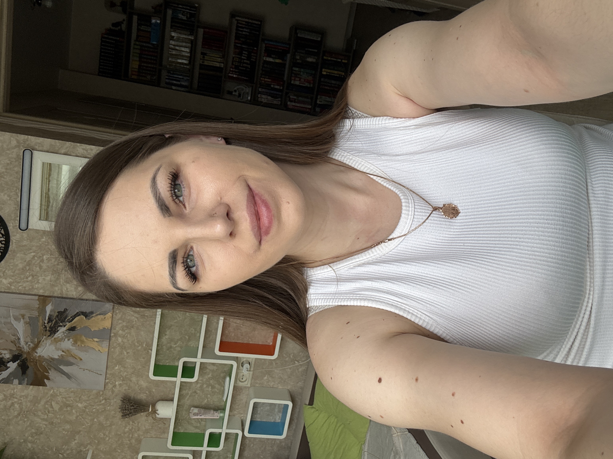 A woman is depicted in a selfie. She has brown hair, fair skin, and is wearing a white ribbed tank top and a delicate gold necklace. She has noticeable eyelashes and is looking directly into the camera with a slight smile. The background is a blurred interior of a room. Behind her are shelves filled with what appear to be books or media. The wall has a patterned wallpaper and an abstract painting, along with some decorative wall elements. The picture appears to have been taken indoors in Раздолье, Russia.