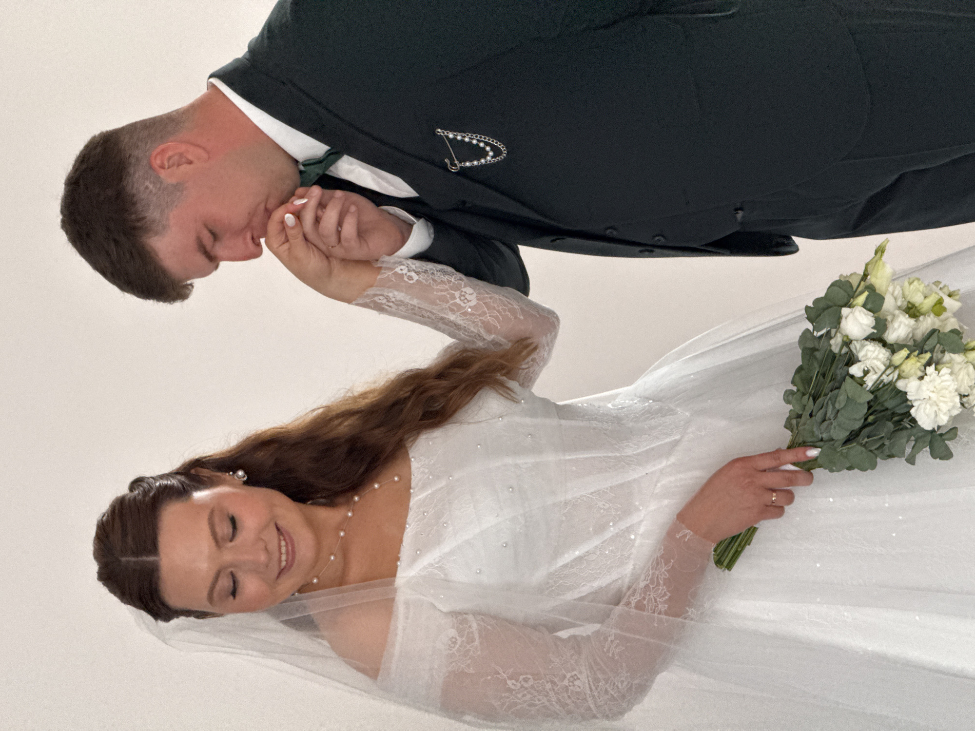 The image, set in Chaikovsky, Russia, features a bride and groom sharing an intimate moment against a plain, light-colored background. The groom, positioned on the left, wears a dark green suit with a white shirt and a dark green tie. A silver brooch, shaped like a 'D' or a stylized 'P' and adorned with small pearl-like beads, is pinned to his lapel. He is gently kissing the back of the bride's hand, his eyes closed, conveying tenderness and affection. His hair is short and neatly styled.
The bride, on the right, is wearing a white wedding gown with long, sheer lace sleeves. Her dress features a translucent overlay with scattered embellishments. She has long, wavy brown hair, a delicate pearl necklace, and pearl earrings. The bride is holding a bouquet of white flowers, including roses and other white blossoms, interspersed with green eucalyptus foliage. Her eyes are closed, and she has a serene, happy smile on her face, reflecting joy and contentment. The overall scene depicts a romantic and emotional connection between the couple, likely captured during their wedding day or a professional photoshoot. There is no visible text in the image.