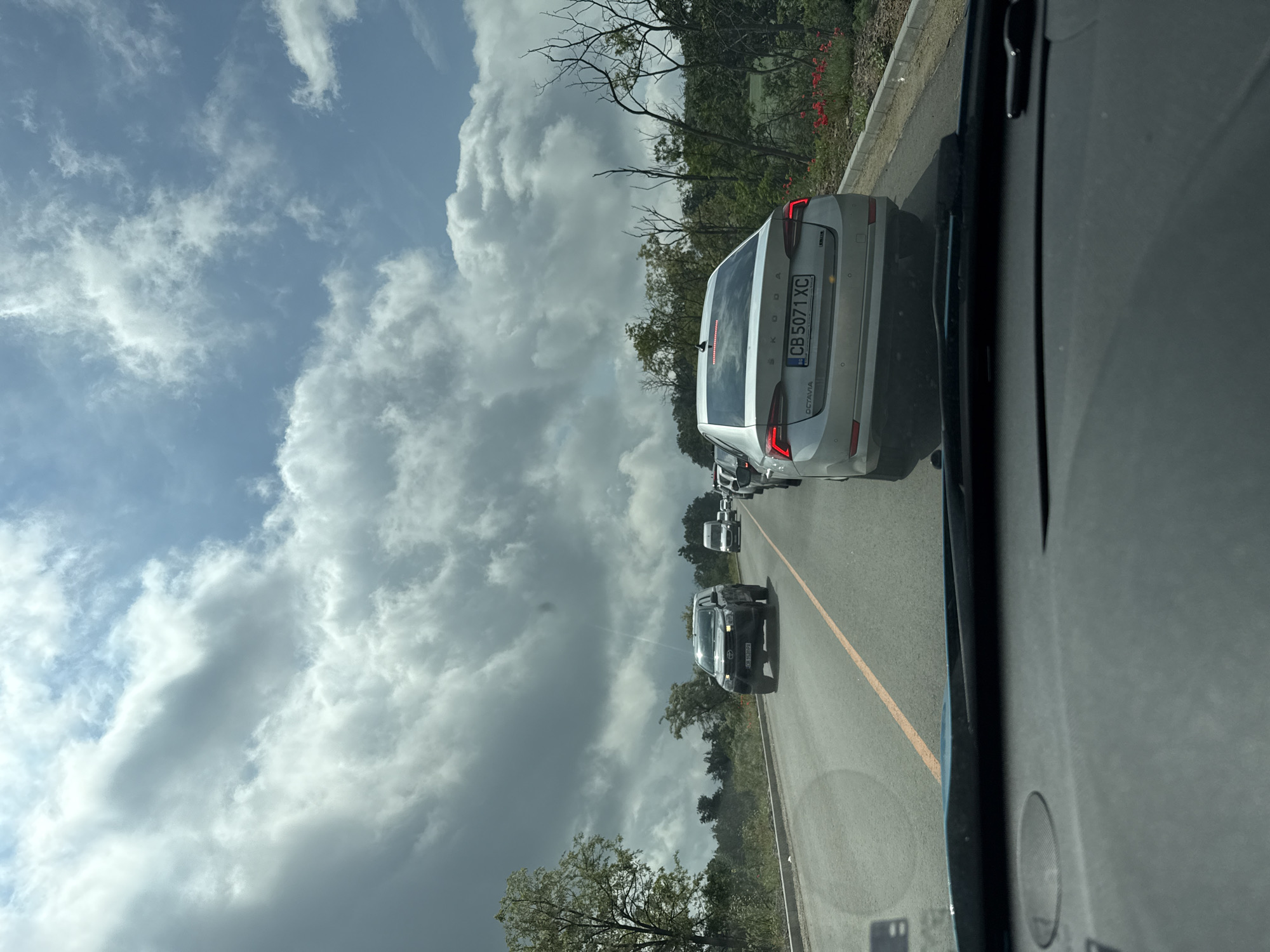 The image, taken from inside a vehicle in Ruen, Bulgaria, depicts a daytime scene on a road under a predominantly cloudy sky. The perspective is looking forward through the windshield, slightly angled.
In the foreground, a silver Skoda Octavia is directly ahead, displaying the Bulgarian license plate "CB 5071 XC." The "SKODA" brand and "OCTAVIA" model names are visible on its rear. Several other cars are lined up further down the asphalt road, suggesting traffic. A dark-colored car with headlights on is particularly noticeable among them. The road features a clear yellow line on what appears to be its right shoulder or edge.
The sky is filled with large, white and grey cumulus clouds, with some areas of blue sky peeking through. Along the left side of the road, there is natural vegetation including various trees, some with green leaves and others with bare branches, interspersed with green bushes and hints of red flowering plants. A portion of the observer's car interior, including a dark dashboard and windshield frame, is visible on the right side of the image. The bright, cloudy conditions suggest a midday setting.