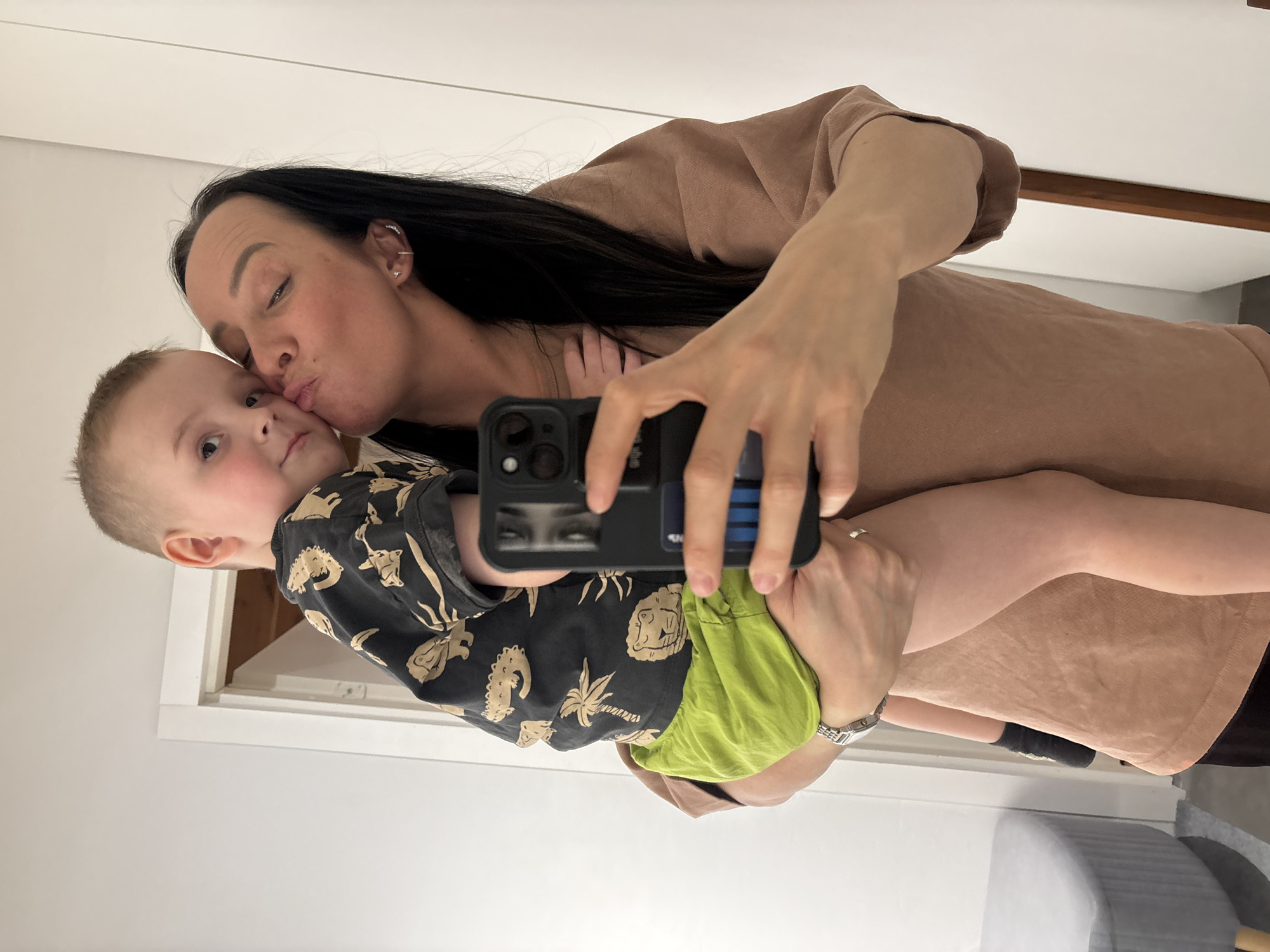 A mirror selfie captures a woman holding a young boy in Chaikovsky, Russia. The woman, with long dark hair, wears a light brown t-shirt, a silver wristwatch on her left wrist, and a silver ring on her right ring finger. She is kissing the boy's left cheek, with her lips pursed. Her right hand holds a black-cased smartphone, revealing its camera lenses and a blue card tucked into the case. The boy, with short light-brown hair, wears a dark short-sleeved shirt with light-colored patterns depicting jungle animals and bright green shorts. He is looking directly at the camera. A white-framed doorway is visible to the left behind the subjects, revealing a darker interior with a wooden ceiling or upper wall. A grey, ribbed upholstered stool is partially visible on the far left. The environment consists predominantly of plain white walls.