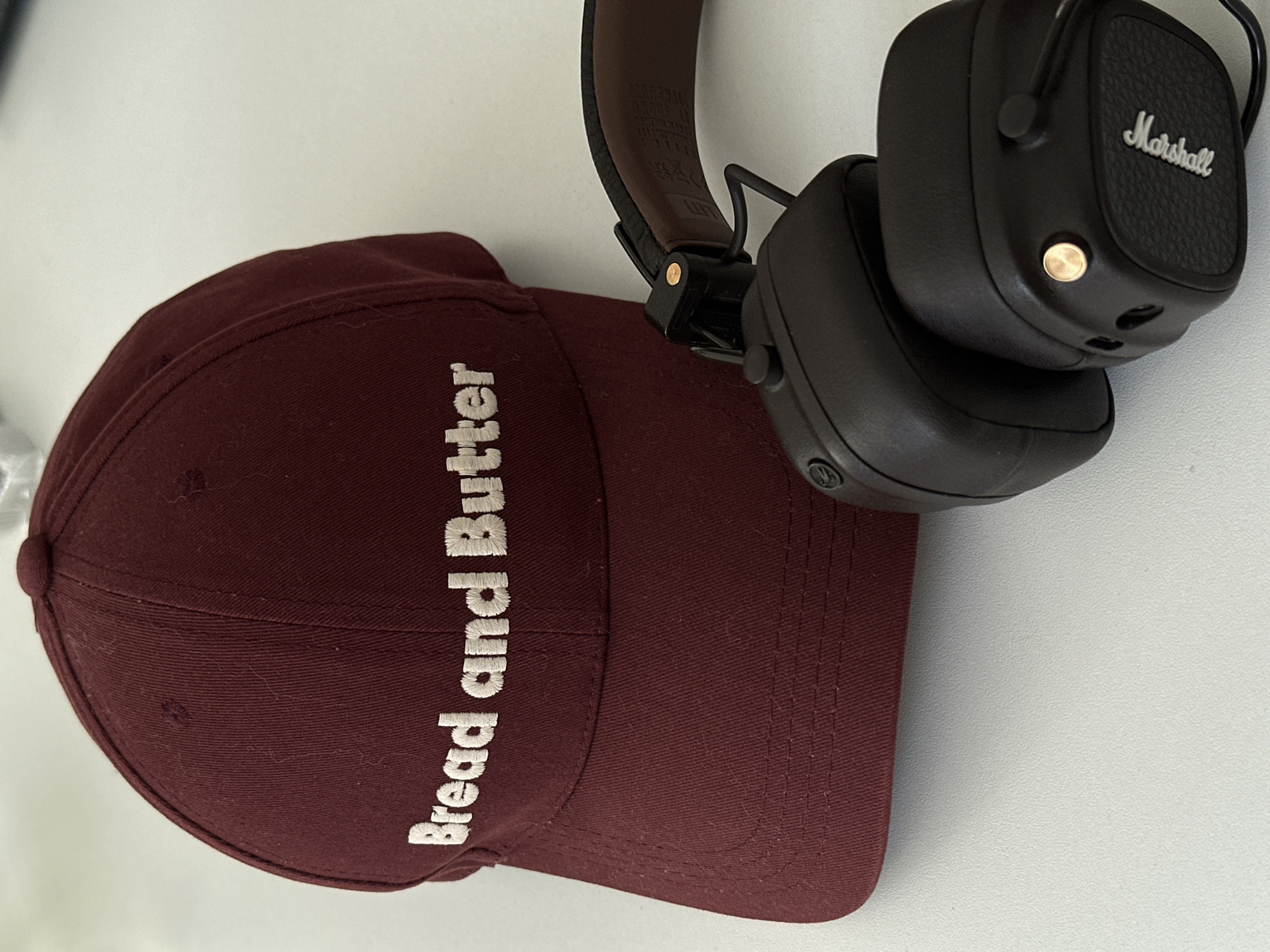 A maroon baseball cap, embroidered with "Bread and Butter" in white text on its front panel, rests on a light-colored, textured surface. To the right and slightly in front of the cap, a pair of black Marshall over-ear headphones are positioned. The headphones feature black leather-like earcups, a visible "Marshall" logo in white on the right earcup, and gold-colored metallic accents, including a control knob on the right earcup. The headband of the headphones also displays a brown, leather-like underside. The background consists of a plain, undifferentiated light color, consistent with an indoor setting. No individuals or specific identifying landmarks are present. The precise location within the city of Moscow, Russia, cannot be determined from the visual evidence in the image.
