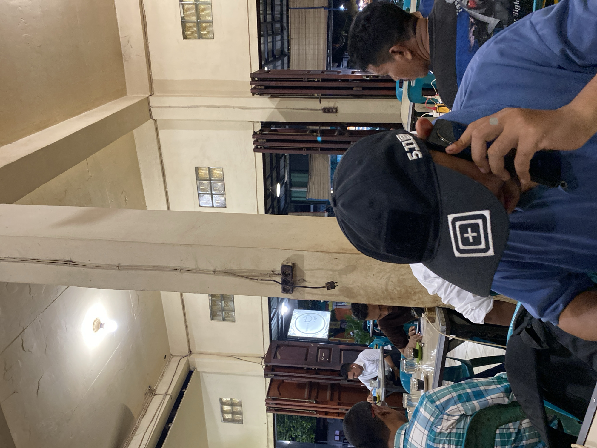 This image depicts a group of people gathered at tables, likely in a restaurant or cafe in Lhokseumawe, Indonesia. The setting appears to be indoors with an open-air feel, as suggested by the visible ceiling, light fixtures, and the presence of what look like shipping containers used as part of the structure. The lighting suggests it is evening or night.
The primary subjects are several individuals, some of whom are partially visible. One person in the foreground is wearing a black baseball cap with a white logo featuring a plus sign inside a square. Another person, in a blue shirt, is holding a mobile phone. Other individuals are seated at tables, some interacting with each other, while others appear to be looking down at their phones or the table. The activity seems to be a casual social gathering, possibly a meal or simply a meet-up.
Notable details include the glass block windows on the walls, the exposed electrical wiring and sockets, and the ambient lighting from ceiling fixtures. The presence of glasses and what might be food or drinks on the tables indicates a dining or social context. The overall mood seems relaxed and informal. Some text is visible on the cap, "5.11," and another logo on a sign in the background appears to be "Kopi." The image is oriented vertically.