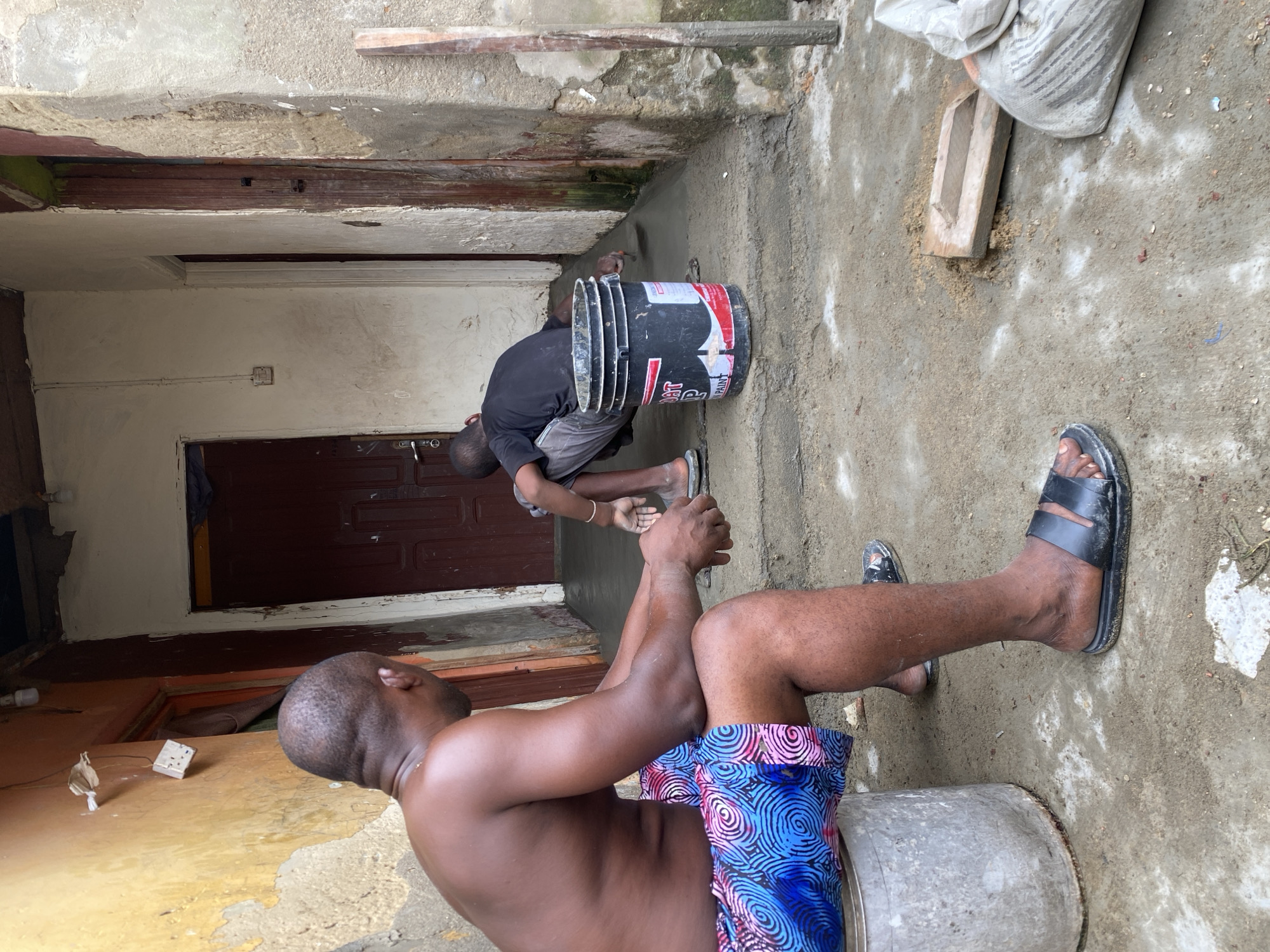 Two individuals are present in an exterior space, likely an alley or courtyard in Ado, Nigeria. One person, shirtless and wearing blue patterned shorts, is seated on an overturned bucket. Another person is bent over, working on the cement floor near a doorway, using a trowel and a paint bucket. A bag of cement and a wooden block are nearby. The walls appear to be made of concrete and are in disrepair.