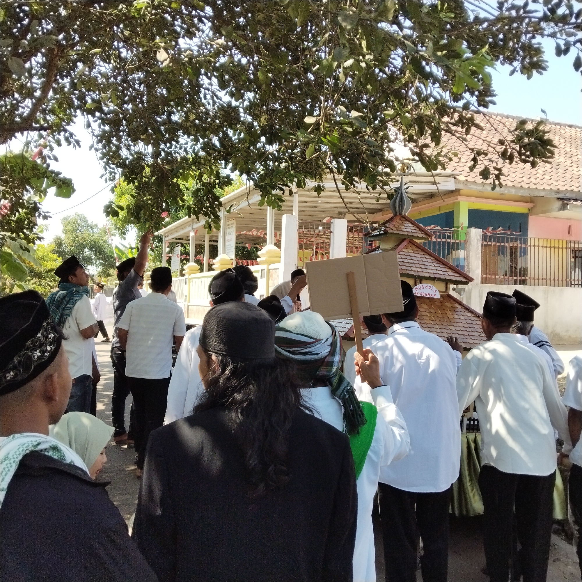 The image depicts a group of men and one woman in what appears to be a street in Pamulihan, Indonesia, with a building resembling a mosque or prayer hall in the background. The scene is set outdoors under a tree with lush green foliage, with a tiled roof building visible behind the people. Several individuals are wearing traditional Islamic attire, including white shirts and black caps (peci). One man in the foreground is holding a cardboard sign. Another man in the middle ground is pointing upwards towards the tree. The building in the background has a multi-colored facade, featuring shades of pink, yellow, and blue, and appears to be a place of worship, indicated by the sign "MUSHOLA AR-ROHMAN" on its roof. The overall atmosphere suggests a gathering or procession.