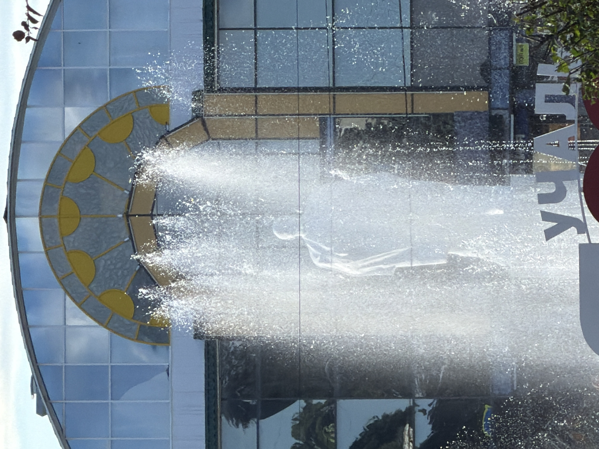 The image captures a dynamic outdoor scene dominated by a large fountain spraying water in the foreground. Behind the fountain, a multi-story building with a curved, glass facade is visible. The glass panels reflect the sky and surrounding environment, with some panels appearing to be tinted blue. An arched section of the building's upper facade features a decorative element with yellow semi-circular shapes set against a translucent, textured material.
A statue, appearing to be a solitary figure in white or light-colored clothing, is partially obscured by the cascading water of the fountain. The statue is positioned in front of the building's facade.
Below the building, a partially visible sign with large, white, block letters can be seen. The visible portion of the sign spells out "УЧАЛ" in Cyrillic script. Green foliage from trees or bushes is present in the lower right corner of the image, partially obscuring the base of the sign. The overall lighting suggests it is daytime, with sunlight creating highlights on the water droplets. The scene is likely an urban public space given the presence of a building, fountain, and signage.