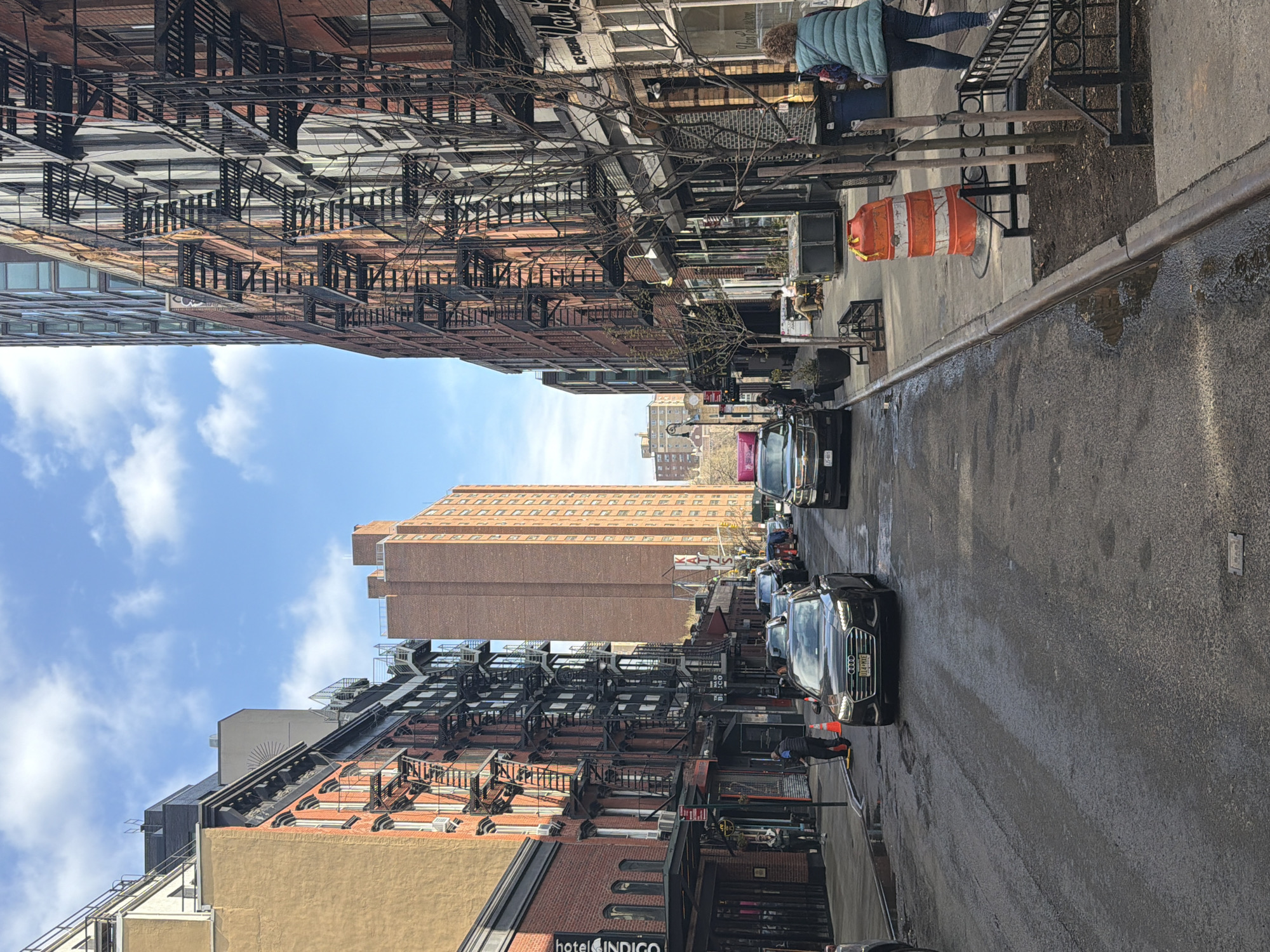 This photograph captures a street scene in New York City, United States. The shot is taken from a slightly elevated position, looking down the street between buildings on either side. The buildings are typical of the city, featuring brick facades and metal fire escapes. A taller building looms in the distance, partially obscured by the surrounding structures. Parked cars line the street, and there's an orange traffic cone visible on the sidewalk. Pedestrians are present on the sidewalks, though they are not the primary focus of the image. The sky above is partly cloudy, indicating a daytime setting. The overall impression is a bustling urban environment.