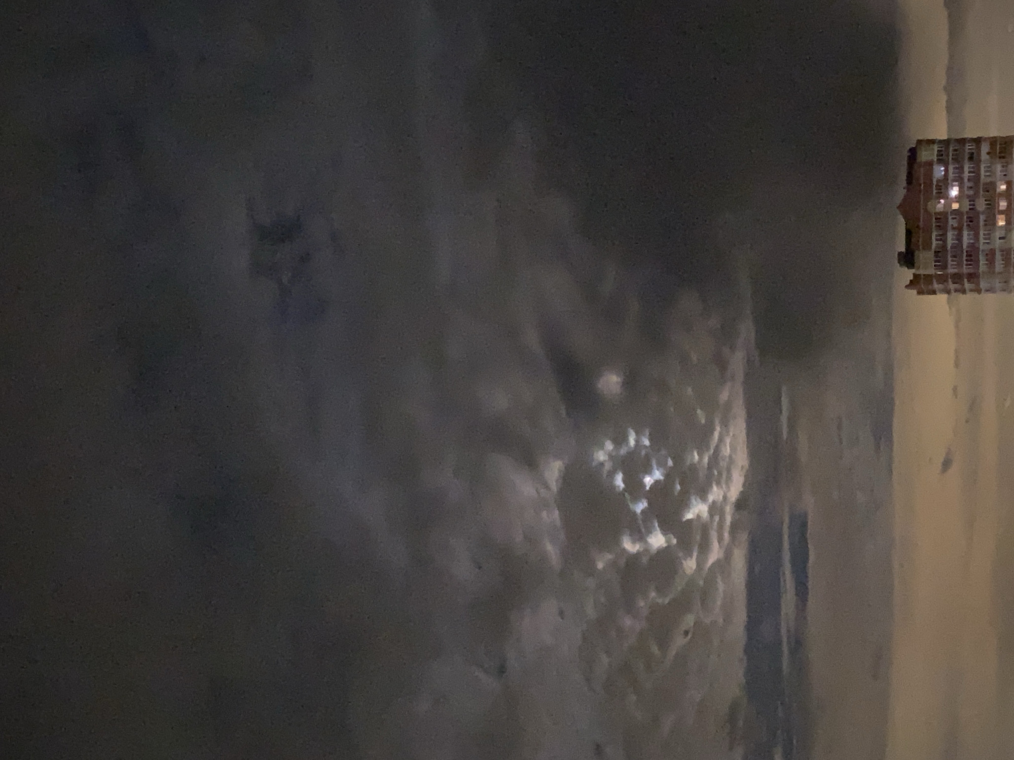 A night-time shot with a significant portion of the frame filled with dark, overcast sky. Light shines through a break in the clouds, creating a bright, amorphous shape. In the upper right corner is a multi-story building with lit windows. The location appears to be a city, but the specific details necessary to ascertain its identity are not present.