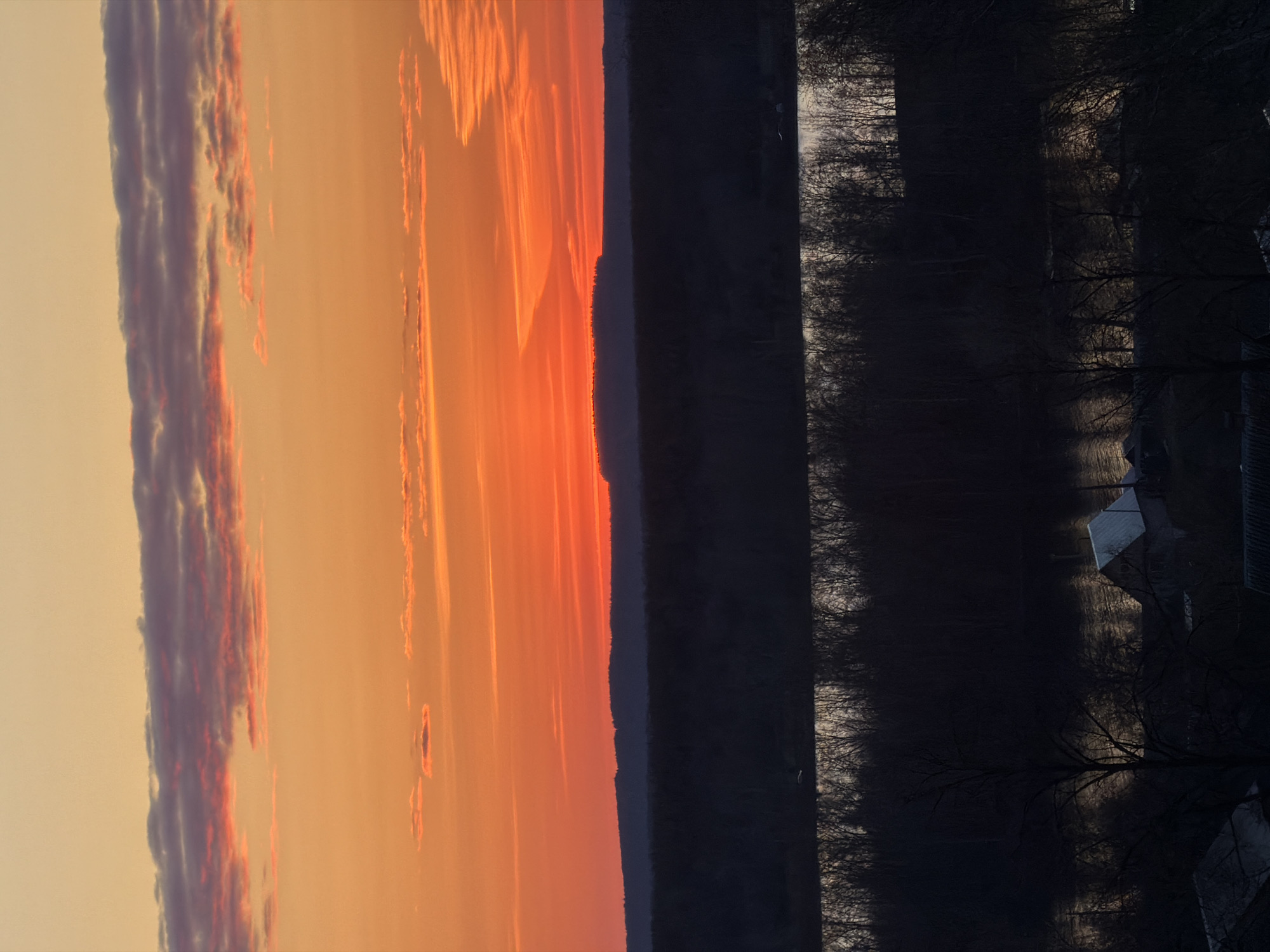 The image shows a landscape at dusk. On the left, the sky transitions from orange to a lighter hue with visible clouds. A dark, undefined mass likely represents a forest or treeline dividing the sky from a darker, shadowed area to the right. Within this darker area, a body of water is discernible, reflecting some light. Bare trees are visible along the water's edge. The rooftops of buildings are also noticeable in the shadowed area, suggesting a residential or built-up environment, possibly in the city of Chaikovsky, Russia.