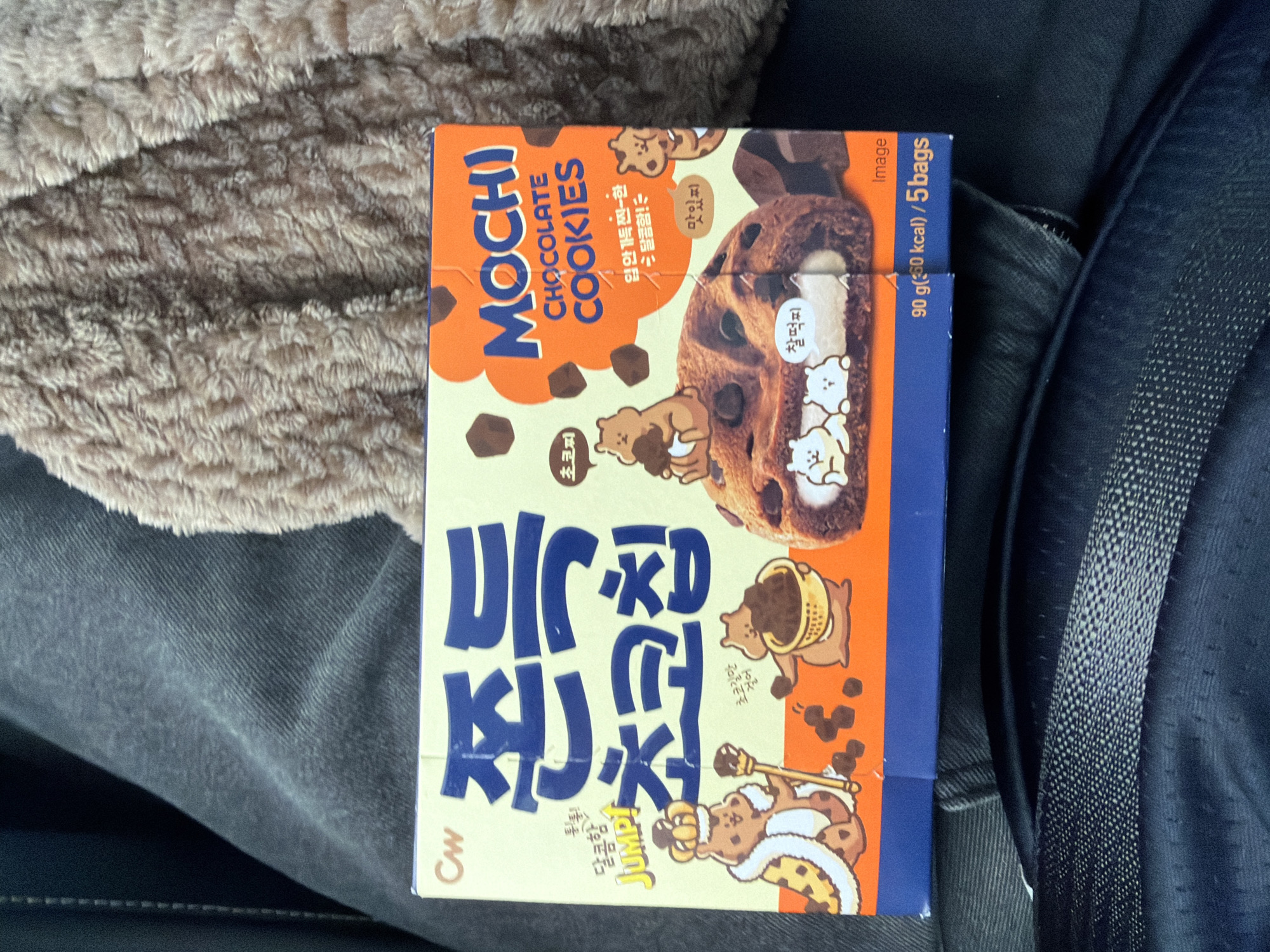 The image shows a close-up of a box of "MOCHI CHOCOLATE COOKIES" by CW. The box is predominantly yellow and orange with blue lettering. It features cartoon illustrations of bears, a large chocolate cookie with mochi filling, and scattered chocolate pieces. Text on the box is in both Korean and English. The box is resting on dark, textured fabric, likely clothing. In the background, a fuzzy, light brown textile is visible. The scene is indoors, possibly in a car given the texture of the fabric, and is not located in Tofta, Sweden. There are no people or discernible actions taking place.