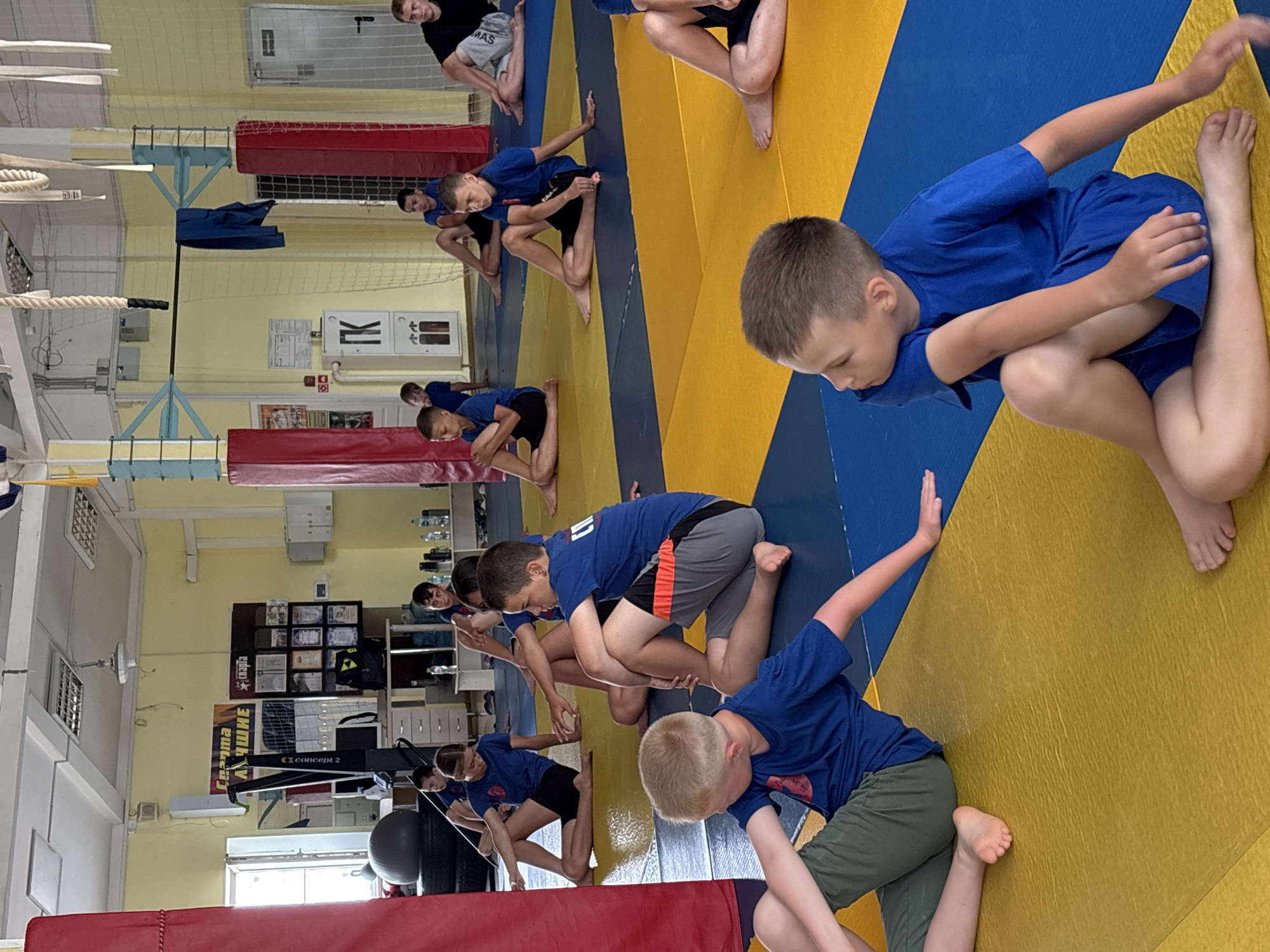 The image depicts a group of young people, predominantly boys, engaged in a stretching or yoga-like activity on a blue and yellow mat in what appears to be a gymnasium. The participants are dressed in athletic attire, mostly blue t-shirts and dark shorts. They are seated on the mat with their legs bent and twisted, suggesting a stretching routine. The background shows a typical gym environment with sports equipment, posters on the wall, and lighting fixtures on the ceiling. A banner with Cyrillic text is visible on the wall, which may indicate the location or the name of the establishment. The overall scene is one of organized physical activity within an indoor sports facility. The poster on the wall with "Герма Ачине" suggests the location is likely a sports club or gymnasium in Chaikovsky, Russia.
