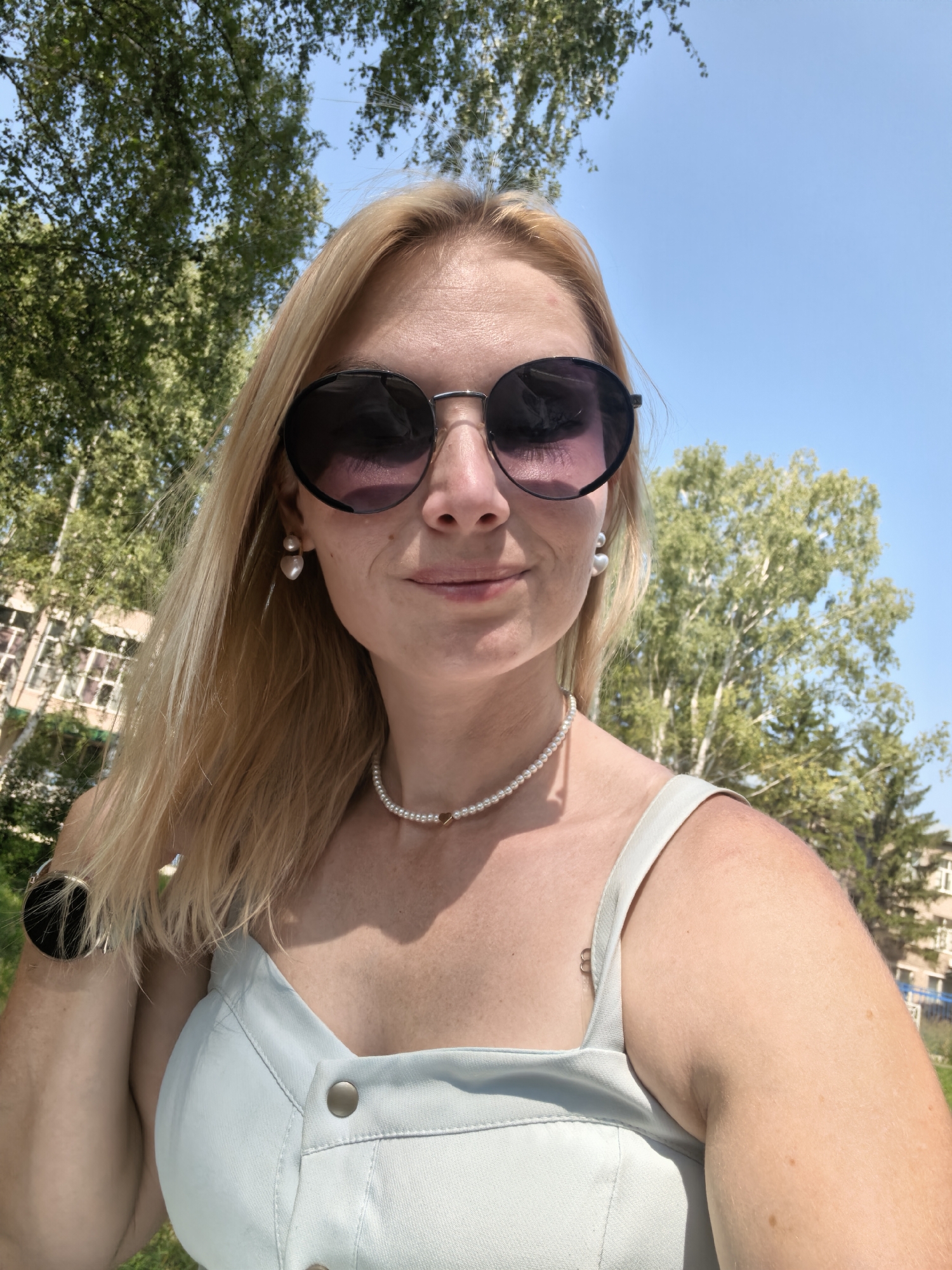 A young woman with blonde hair and sunglasses takes a selfie outdoors. She is wearing a light blue dress and pearl jewelry, including earrings and a necklace with a small gold heart pendant. The background shows green trees and a blue sky. The image is cropped to focus on the woman's face and upper body.