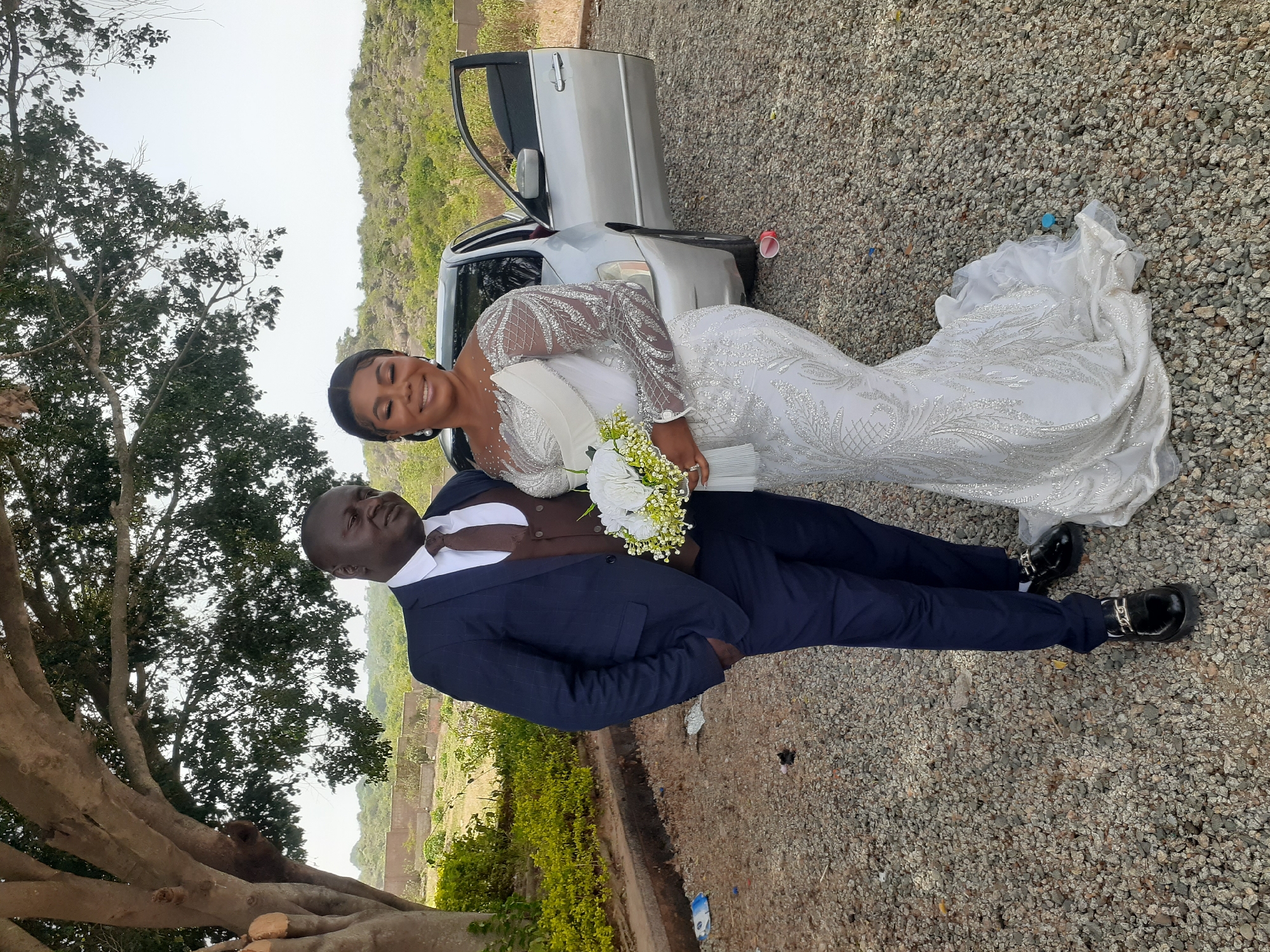 The image depicts a wedding couple in an outdoor setting, likely in the city of Jos, Nigeria. A woman in a white, long-sleeved, sequined wedding gown reclines against a man dressed in a navy blue suit with a brown vest and tie. The woman is holding a bouquet of white flowers and smiling. They are positioned near a silver car with an open door. The ground is covered with gravel. The background includes a tree, low vegetation, and a hillside.
