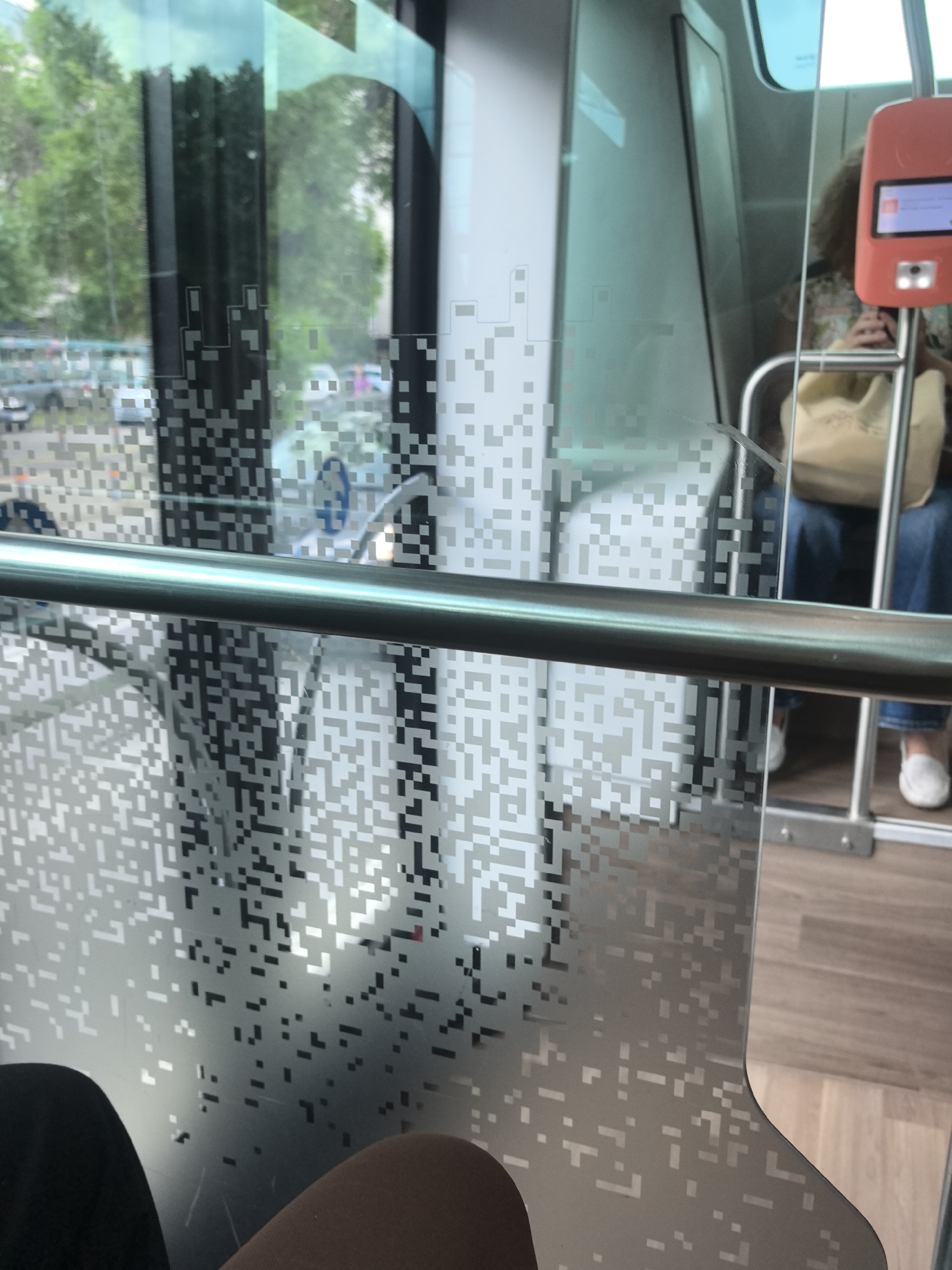 The image shows the interior of a public transportation vehicle, likely a tram or light rail, based on the seating and window design. A stainless-steel grab rail is prominently in the foreground. The window is patterned with a dense, pixelated design that obscures the view outside, where trees and other vehicles are visible. Seated behind the glass barrier is a person with blonde, curly hair. She is wearing a light-colored floral top, blue jeans, and white slip-on shoes. She holds a tan purse on her lap. An orange ticket validation machine is affixed to the frame. Another seated passenger is partially visible in the bottom of the frame, displaying black and tan clothing. The interior floor is made of light brown wooden planks.