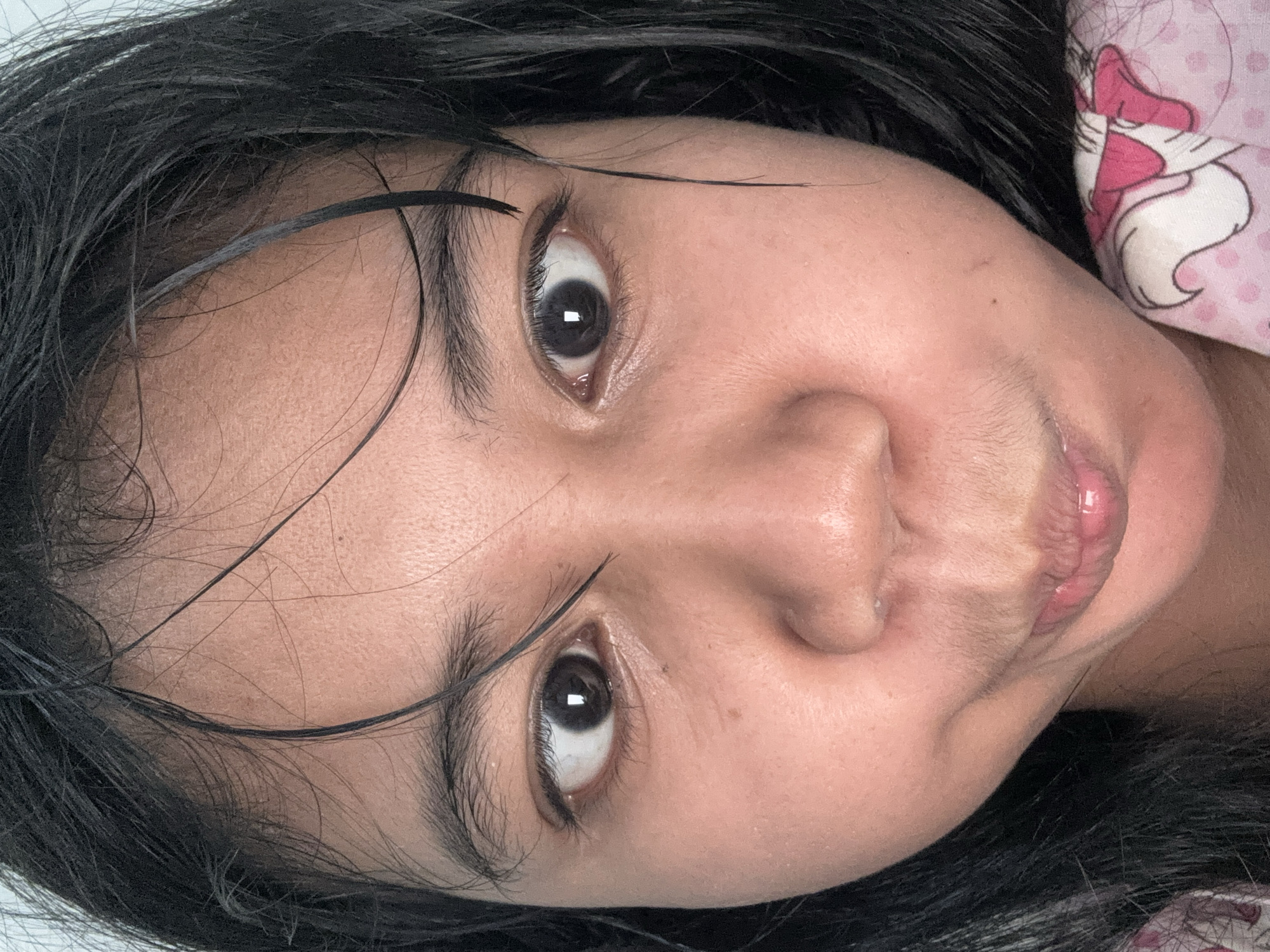 A close-up, slightly angled, indoor photograph features the face of a young East Asian individual. The subject's eyes are wide open and looking directly at the camera, with their mouth pursed into a playful expression. Their dark hair is wet and sticks to their forehead and cheeks. The lighting suggests an indoor setting, possibly a home environment, with a hint of patterned fabric, likely clothing or bedding, visible in the lower right corner. The photograph's composition and the subject's expression suggest a candid or playful moment. No specific location in North Jakarta, Indonesia, can be determined from the visual information provided.