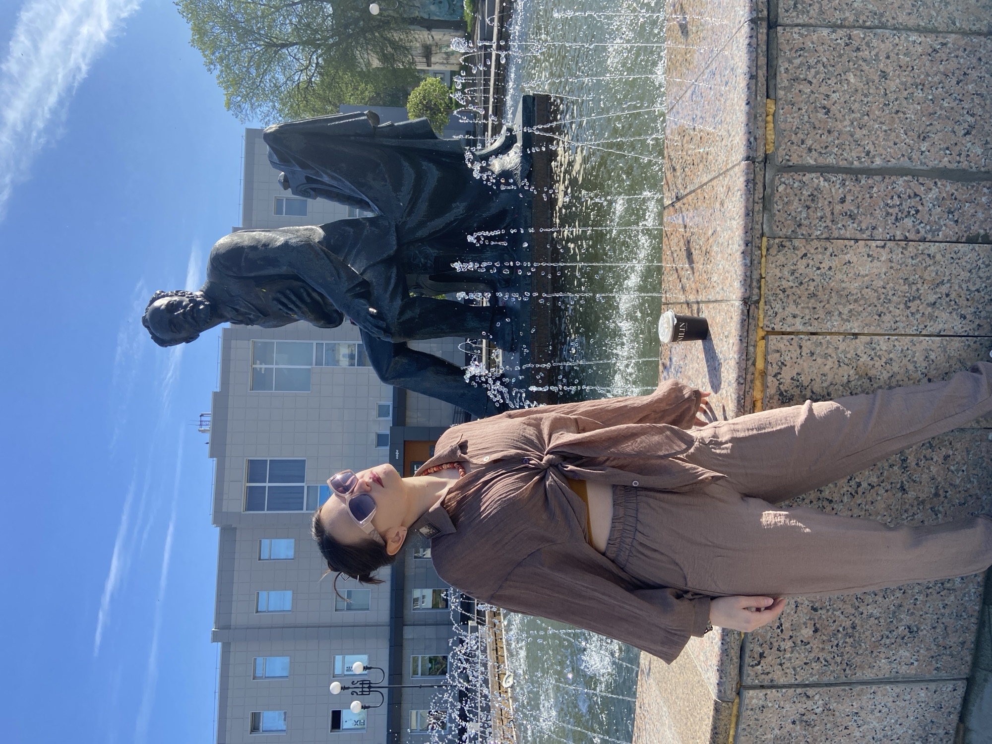 A woman is reclining against the edge of a fountain in Chaikovsky, Russia. She wears brown sunglasses and a brown outfit, consisting of a button-down shirt tied at the waist and loose-fitting pants. Her head is tilted back, facing the sky. Behind her is a statue of a man, draped in what appears to be a cloak, standing in the fountain. Water is visibly spraying from the fountain. In the background, there is a modern building with multiple windows and street lamps. The sky is blue with light clouds. A can of "Nulin" is visible on the fountain ledge.