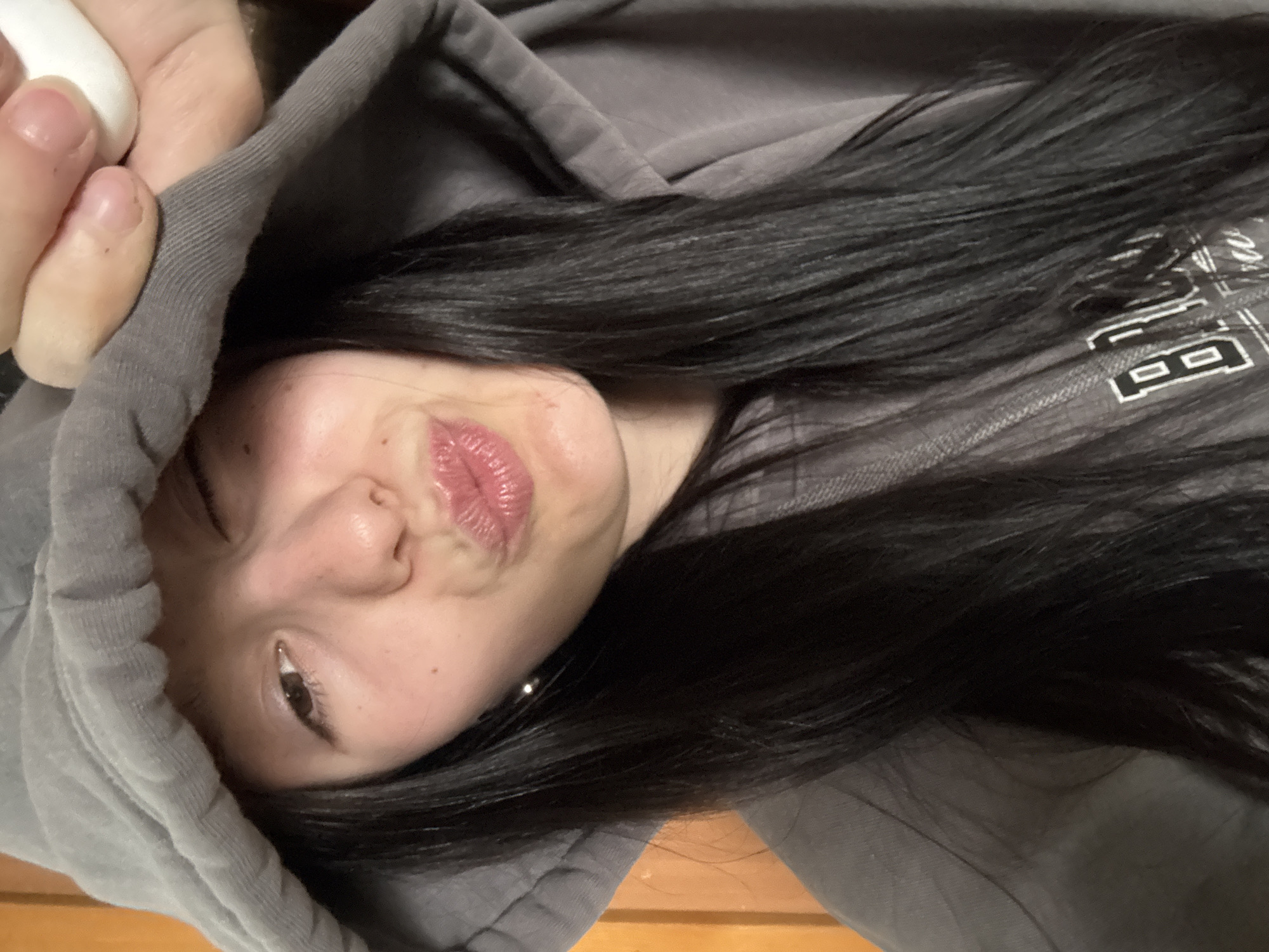 A close-up, tilted portrait features a person, likely female, with long, straight dark hair, looking towards the viewer. The person is wearing a gray hooded sweatshirt, with the hood partially framing their face. Their lips are pursed in a "kissing" or "duck face" expression, highlighted by glossy pink lipstick. One dark eye is clearly visible.
At the top left of the image, a hand with short, neat fingernails is visible, holding a small white object, possibly an earbud or a piece of fabric. The person's dark hair falls over their shoulder and across the front of the hoodie. White text is partially visible on the chest area of the hoodie, with the letter "B" clearly distinguishable, followed by other indistinct letters or symbols. The background appears to be an indoor setting, with a blurred wooden surface visible at the bottom right. The overall impression is a casual and playful moment, possibly a selfie.