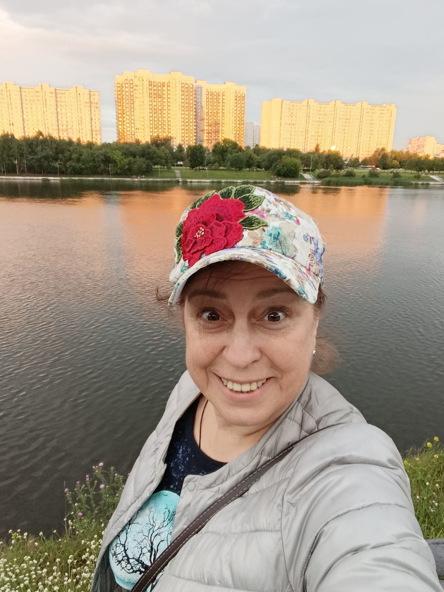 A woman is taking a selfie by a body of water with a cityscape in the background. The woman is wearing a floral cap with a large red flower and a light-colored puffer jacket. Her expression is joyful, with wide eyes and a bright smile, revealing her teeth. She has a dark t-shirt on with a graphic of a tree. The background shows several tall, modern apartment buildings reflecting in the water, suggesting an urban setting. The sky appears to be overcast with the warm glow of sunset, indicating it is likely evening. The foreground features a grassy embankment with small white and pink flowers. The water is calm with gentle ripples. The scene suggests a relaxed moment outdoors, possibly in a park or along a waterfront promenade in Moscow, Russia, during a pleasant time of day. There is no visible text in the image.