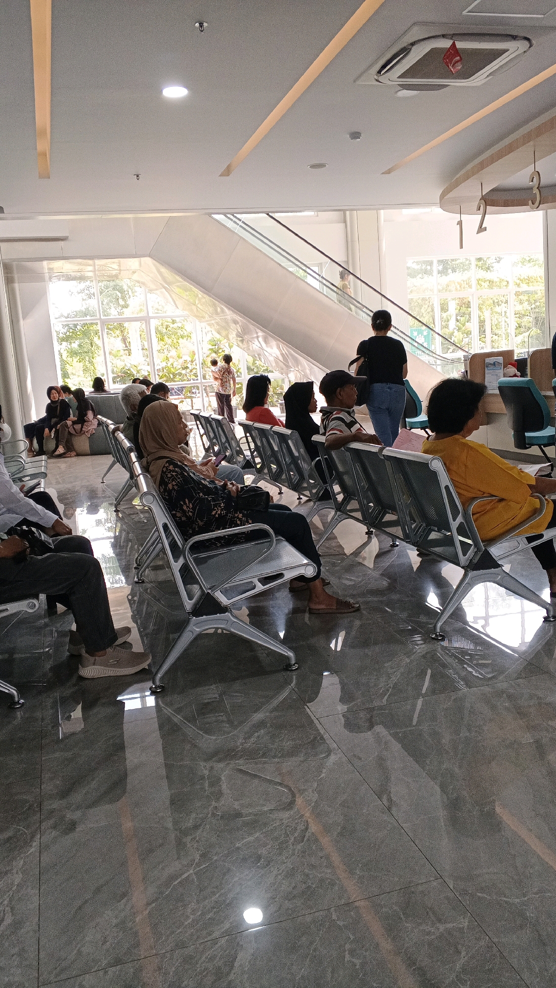 This image depicts the interior of a modern waiting area, likely a public building such as a hospital or government office, in Semarang, Indonesia. The space features a polished grey marble floor that reflects the overhead lighting and the surroundings. Rows of silver-colored metal chairs with dark seats and backs are arranged for seating. Natural light streams in from large windows that look out onto greenery and trees, suggesting a daytime scene. An escalator is visible in the background, indicating a multi-level building.
Several individuals are seated and waiting. In the foreground, a person in dark pants and a light-colored long-sleeved shirt is seated on the left, their feet visible in light-colored sneakers. Further back, a woman wearing a patterned long-sleeved top and a hijab is seated, looking at her phone. To her right, another woman in a black hijab is seated. A man wearing a striped shirt and a cap is seated in the middle-right. On the far right, a woman in a bright yellow long-sleeved shirt is seated.
In the mid-ground, a woman in a black shirt and blue jeans stands facing away from the camera, with a bag slung over her shoulder. Behind her, a man in a patterned shirt is standing and looking towards the escalator. Several other people are visible seated in the background, some with hijabs. The overall atmosphere is one of quiet waiting. The lighting suggests it could be morning or afternoon. The numbers "1", "2" are visible on a curved structure in the upper right, potentially indicating service counters or zones.