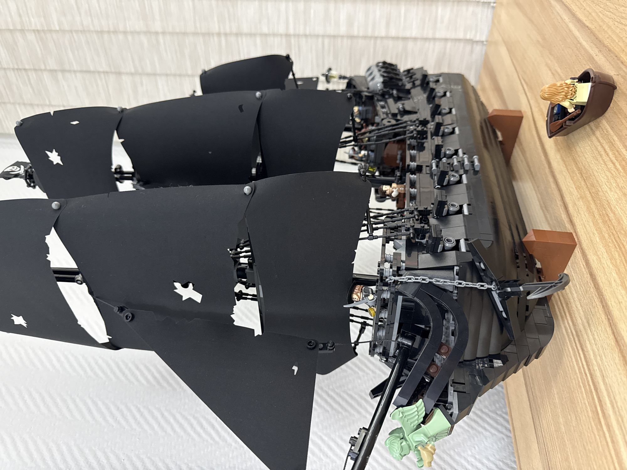 A Lego model of the Black Pearl ship is positioned on a horizontal surface, which is likely a table. The ship is constructed predominantly of black Lego bricks, with brown accents visible in the hull. The sails are black and torn. The ship is docked beside a small rowboat attached to the shelf with a figure seated inside. The background consists of white blinds and a light wood wall. The image does not contain any discernible information about the location being in the city of Krasnoyarsk, Russia.