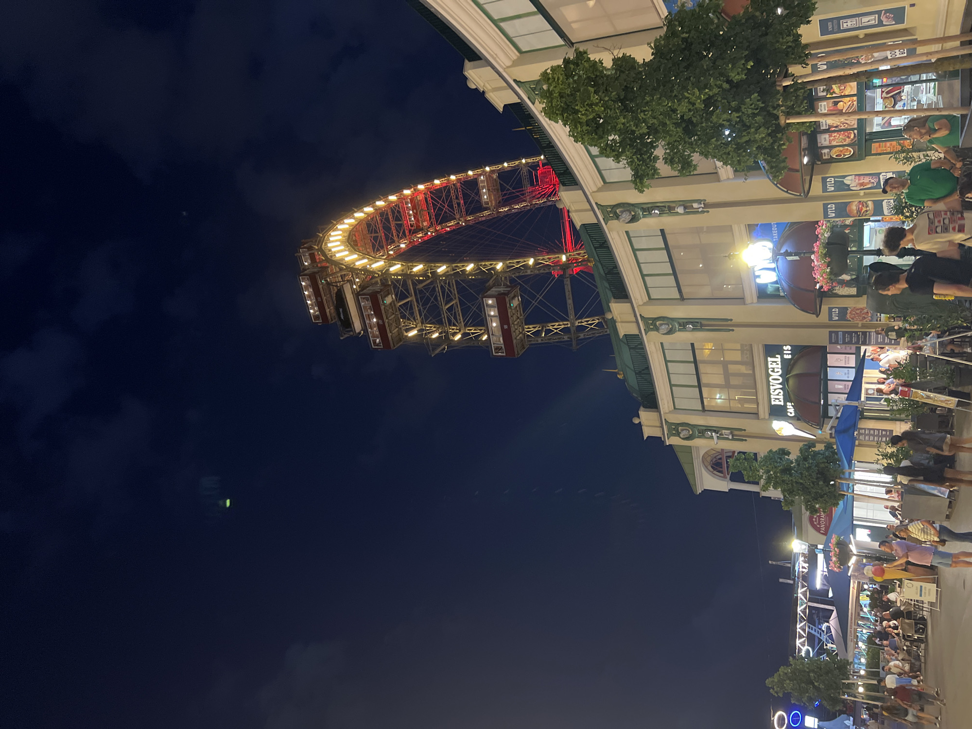The image shows a night scene at the Prater amusement park in Vienna, Austria. The iconic Wiener Riesenrad, a large Ferris wheel, is illuminated with red and yellow lights and dominates the upper portion of the frame. In the foreground, there is a street lined with buildings housing shops and restaurants. People are walking along the street, some are sitting at outdoor tables. The sky is dark blue with some clouds. The overall atmosphere is lively and festive.