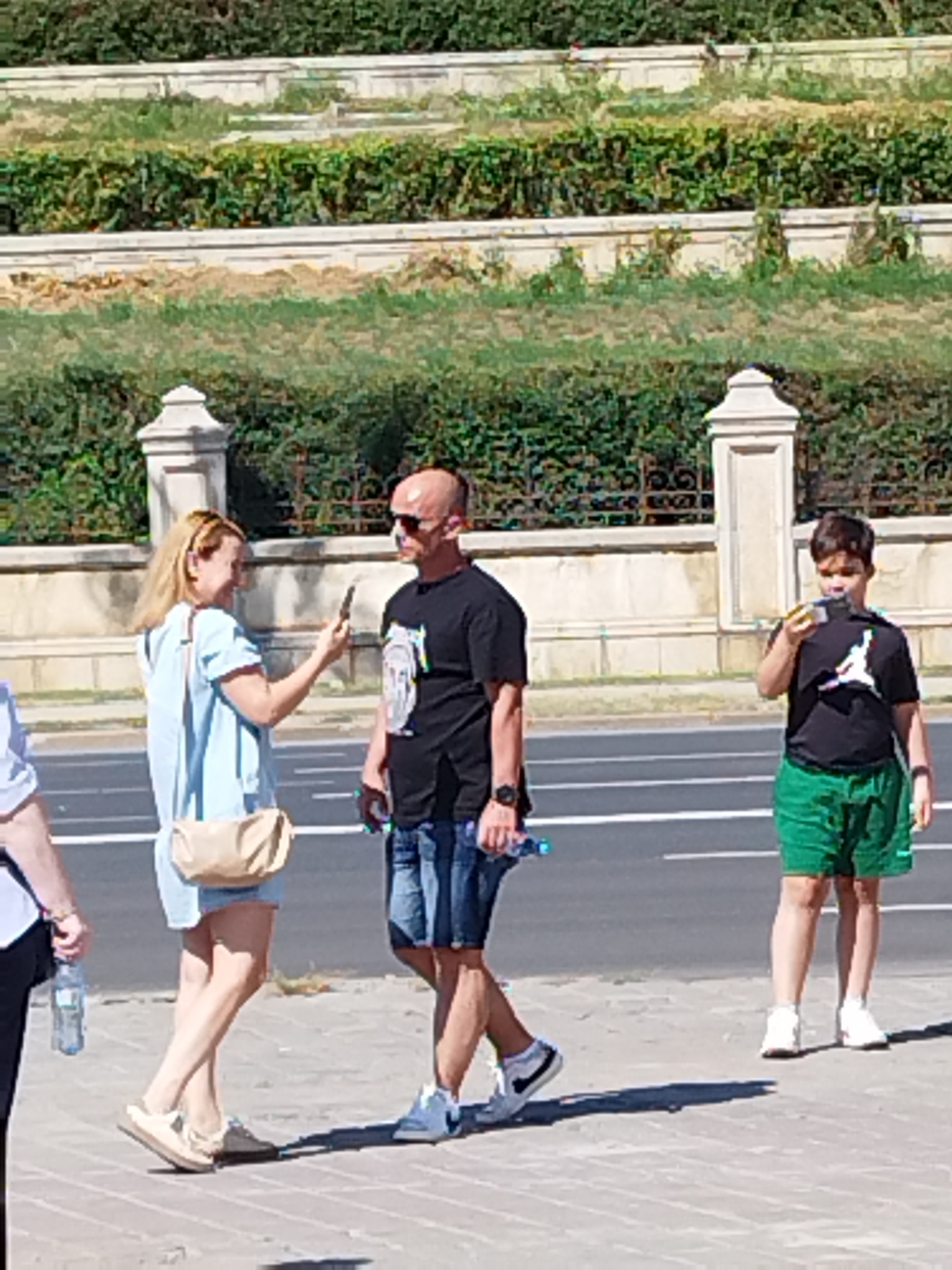 Three people, two adults and one child, are standing on a sidewalk next to a road. In the foreground, a woman wearing a light blue dress and a beige purse is looking at her phone. To her right, a bald man in a black t-shirt and denim shorts is also looking at his phone. To the right of the man, a child in a black t-shirt with a white Jordan logo and green shorts is holding a small object to his mouth, possibly eating. The background shows a tiered, landscaped area with greenery and stone walls, typical of Bucharest, Romania. The road has white lane markings.