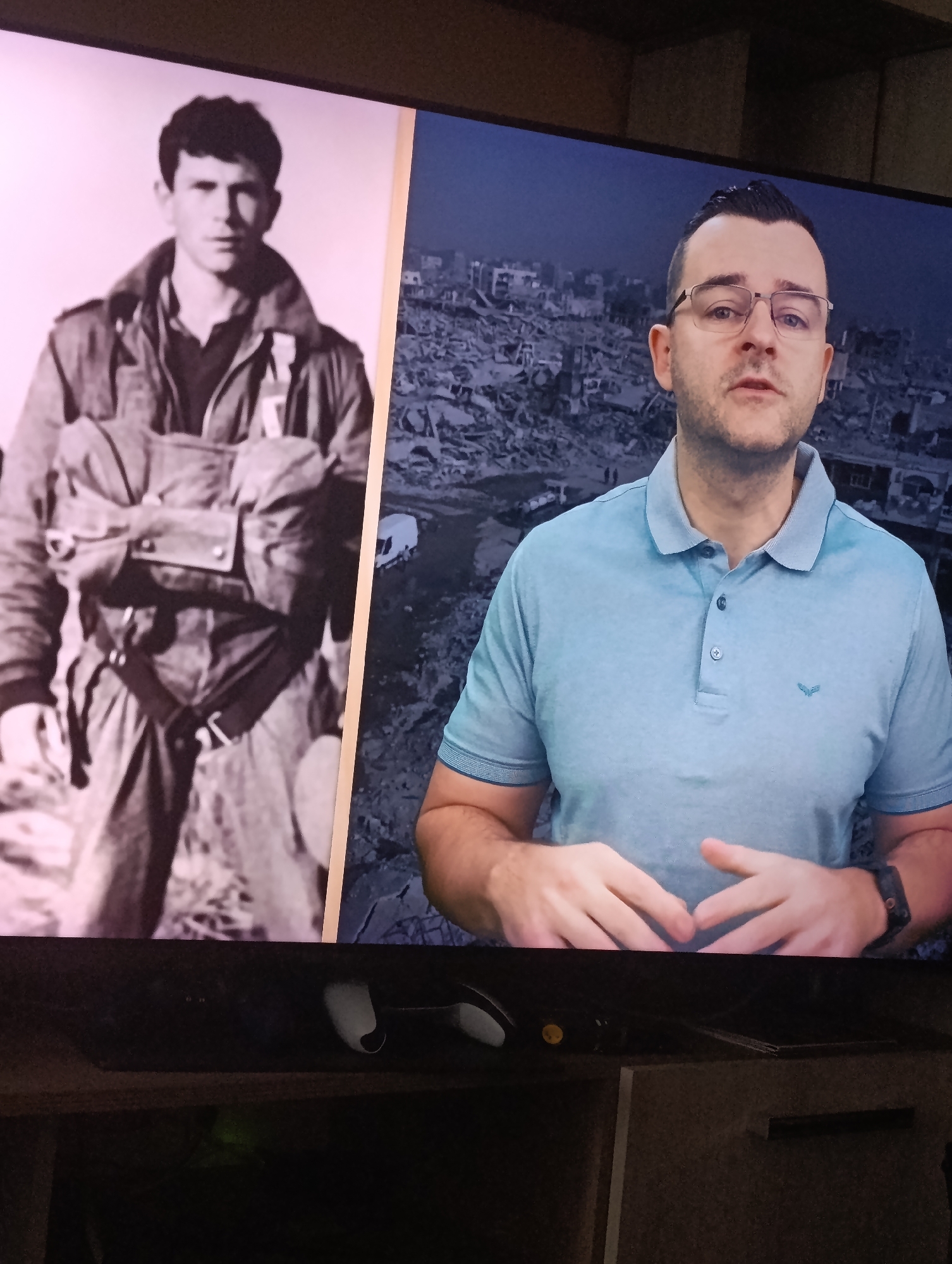 The image shows a television screen displaying two images side-by-side. On the left is a black and white photograph of a young man wearing what appears to be a military or pilot's uniform with a parachute or harness. He has short, dark hair and a serious expression. On the right is a color image of a man wearing glasses and a light blue polo shirt. Behind him is a blurred, grayscale image of a devastated urban landscape, suggesting a war zone or area of significant destruction. There is no indication of location within Plovdiv, Bulgaria. Below the television screen is a shelf unit holding what appears to be gaming consoles or other electronic devices.