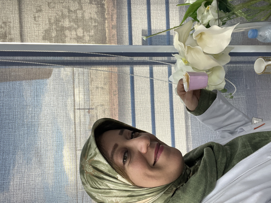 A woman in a green floral headscarf holds a small, light-purple cup. She wears a white lab coat over a green shirt. White calla lilies are arranged in the background, along with a water bottle and a teacup. Light streams through window blinds, casting striped shadows on the wall behind her.
