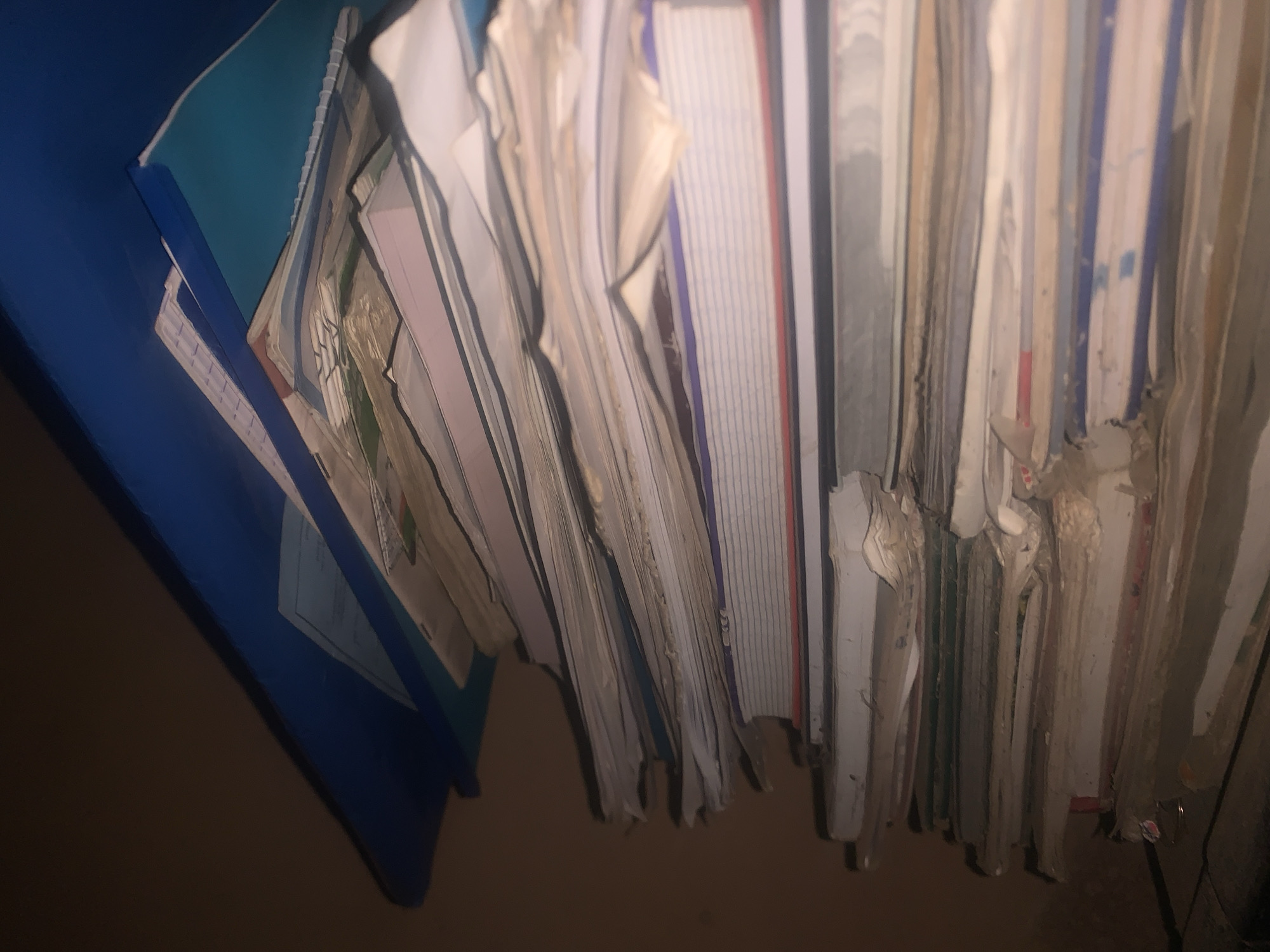 The image shows a close-up, low-angle view of a tall stack of papers and notebooks on a shelf. The items are mostly white and cream-colored, with some showing faint lines or colored covers. The stack is leaning slightly to the right. At the top of the stack, there is a blue binder or folder, and another teal-colored notebook or folder is partially visible underneath it. The background is dark and appears to be a wall. The lighting is dim, casting shadows and highlighting the textures of the paper. There are no people or visible activity in the image. The location in Jos, Nigeria is not discernible from the visual information provided in the image.