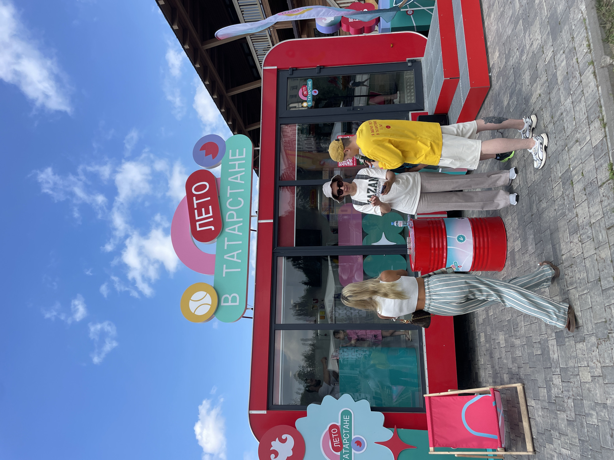 The image depicts an outdoor scene in front of a bright red kiosk with a large sign overhead that reads "ЛЕТО В ТАТАРСТАНЕ" (Summer in Tatarstan). The kiosk has large glass windows and a red staircase leading up to the entrance. To the left, a red and pink beach chair is set up on a grey brick-paved ground. To the right, two red cylindrical objects, resembling barrels, are stacked.
In the foreground, a woman with blonde hair, wearing a white crop top and striped pants, walks away from the camera. She is carrying a brown purse. In the center, a woman wearing a white bucket hat, sunglasses, and a white t-shirt with the word "KAZAN" printed on it, is speaking to a man. She is holding a small item in her hands. The man, wearing a yellow t-shirt and white shorts, is facing the woman and appears to be listening. He has a tattoo on his left calf.
Inside the kiosk, several people are visible, including a man behind a counter and a young girl. The environment appears to be a public space with trees and a building with a wooden roof visible in the background. The sky is blue with scattered white clouds. The overall scene suggests an event or promotional activity related to summer in Tatarstan, likely in Kazan, Russia, given the signage.