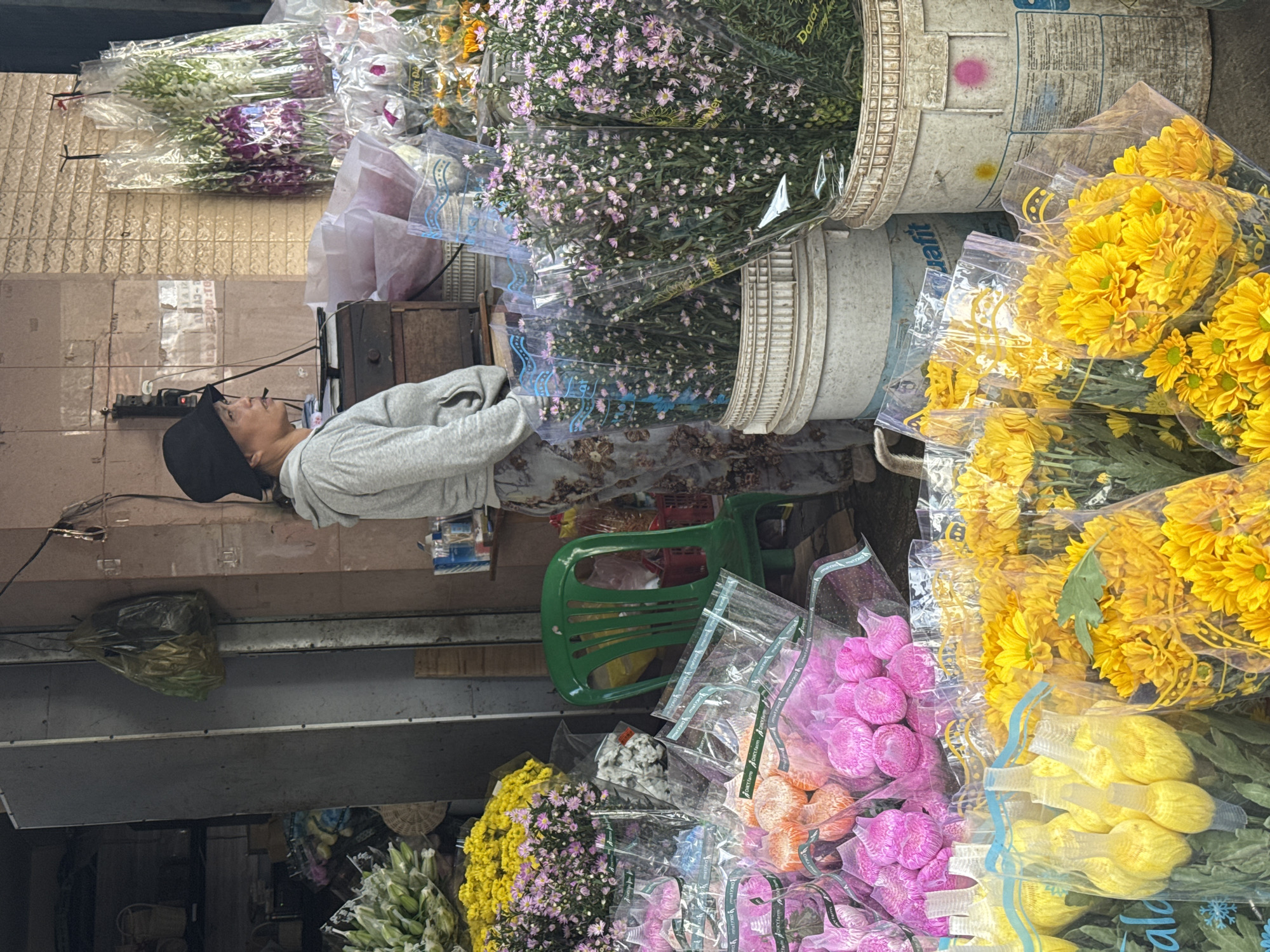 A middle-aged Vietnamese woman wearing a gray hoodie and a black hat stands in front of a flower stall in Nha Trang, Vietnam. She is looking to her left, with a cigarette in her mouth. The stall is filled with various flowers, including yellow chrysanthemums, pink flowers, and white and purple orchids. Some of the flowers are displayed in buckets, while others are wrapped in plastic. A green plastic chair is placed behind the woman. The stall appears to be located on a street, with a building in the background. The overall impression is of a bustling market scene, with the woman as a central figure amidst the colorful display of flowers.