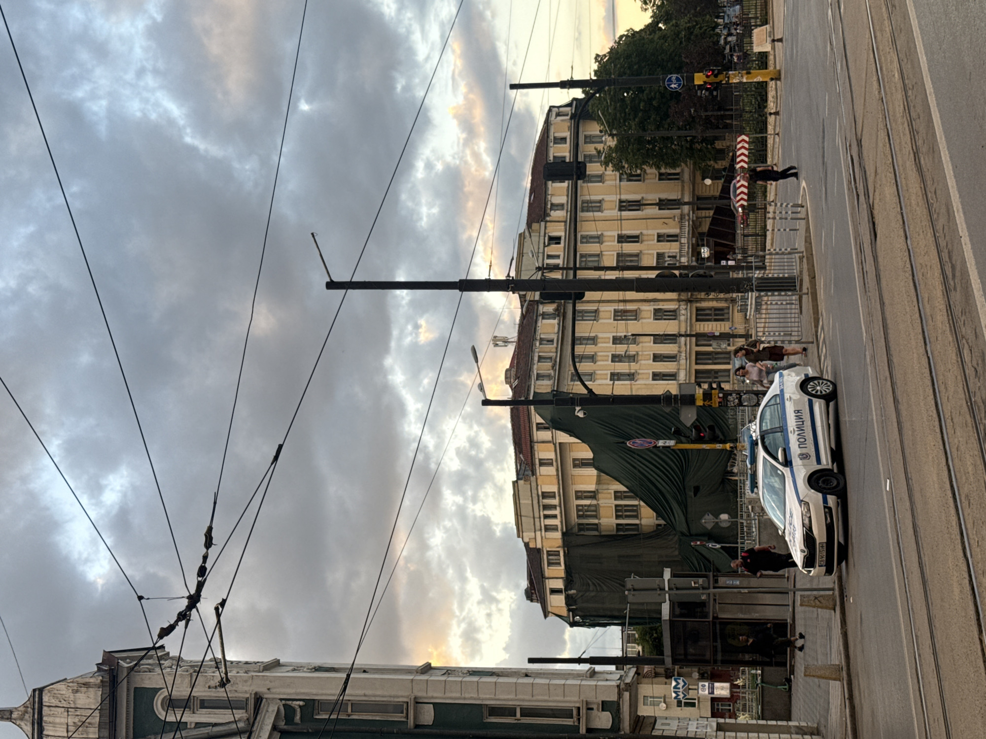 This is a vertical, eye-level, medium shot of a city street scene in Sofia, Bulgaria. The sky is overcast with dramatic clouds. In the foreground, there are tram tracks on the street. A white police car with blue and white markings is parked on the right side of the street, facing left. The car has "ПОЛИЦИЯ" (Police) written on the side in Cyrillic. A traffic light is visible above and to the right of the police car. In the middle ground, a large, yellow building with many windows is under construction or renovation, covered in a dark green tarp. Several people are walking on the sidewalk in front of this building, and a few more are visible across the street. A street lamp stands in front of the yellow building. To the left of the yellow building, a portion of a taller, white and green building with arched windows is visible. Power lines crisscross the sky throughout the image. The scene appears to be a busy urban intersection.