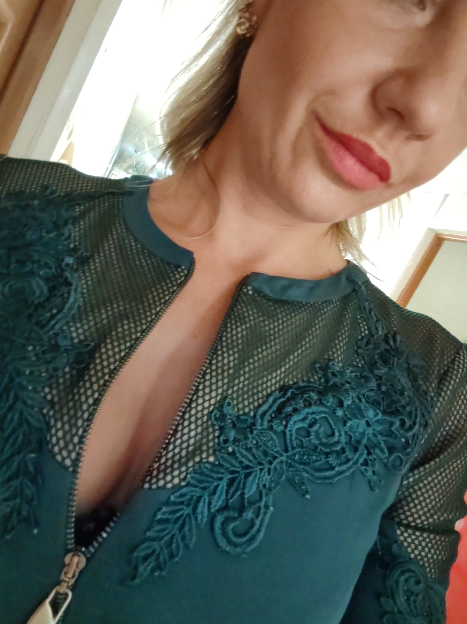 The image is a close-up, waist-up selfie of a fair-skinned woman with blonde hair, taken from a low angle. She is wearing a dark teal dress with lace detailing and a zippered neckline that is partially unzipped, revealing her cleavage. Her makeup includes red lipstick and what appears to be an earring in her left ear. The background is out of focus and appears to be an indoor setting with a bright light source, possibly a window or a light fixture, casting a strong glare. The overall lighting is harsh.