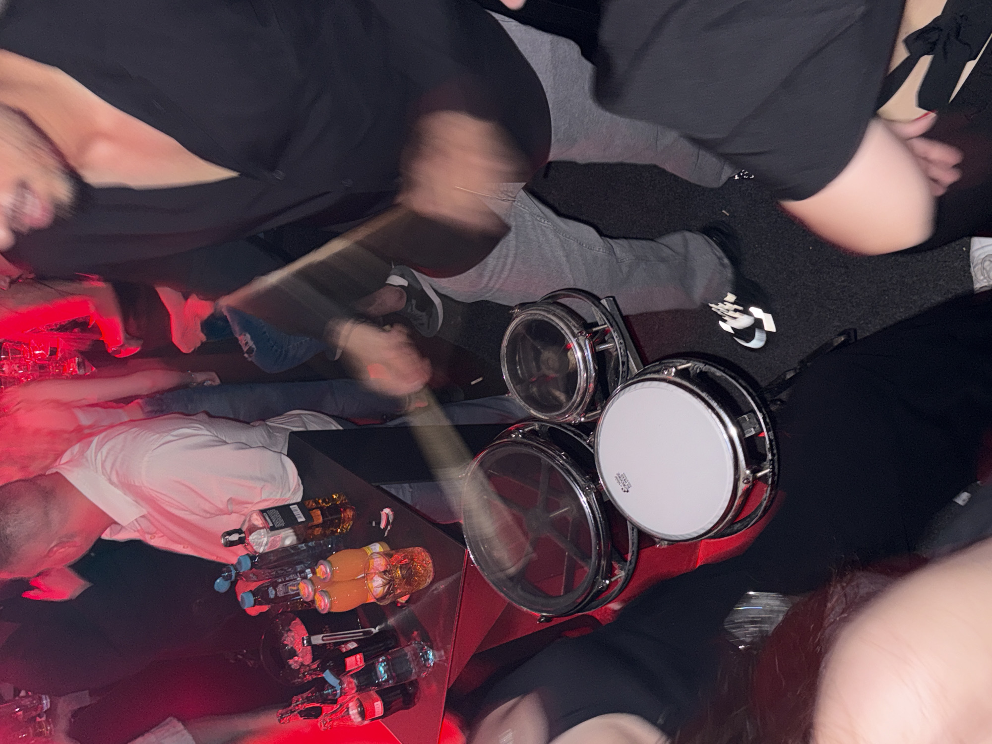 A low-angle shot shows a person playing drums on a red, glass-topped table, possibly in a crowded bar. The drums are a set of three, with chrome hardware. A drumstick is in motion, striking one of the drums. The table holds multiple bottles of liquor, soft drinks, and juice. Several people are gathered around, including one wearing a white shirt and another in a black outfit. The environment appears dimly lit with a red hue. The location is likely in Sofia, Bulgaria.