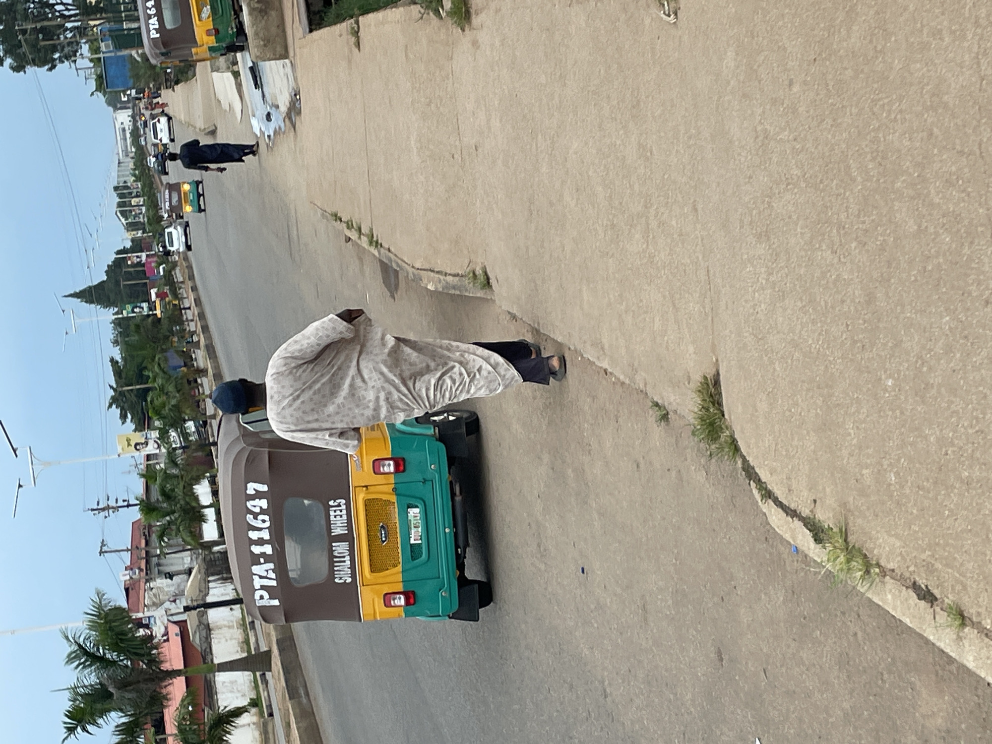 A man in a white patterned robe and a dark blue cap walks away from the camera on a concrete sidewalk. He is walking on the right side of the road, which has a beige asphalt surface. In front of him, to his left, is a three-wheeled auto-rickshaw, predominantly yellow and teal, with the number "PTA-11647" visible on its brown rear canopy. The rickshaw also has "SHALLOW WHEELS" written on its back. Behind the rickshaw, there are other vehicles and a few pedestrians visible on the road. The environment appears to be a street scene in Barakin Lerawi, Nigeria, with palm trees and buildings lining the road. The sky is overcast.