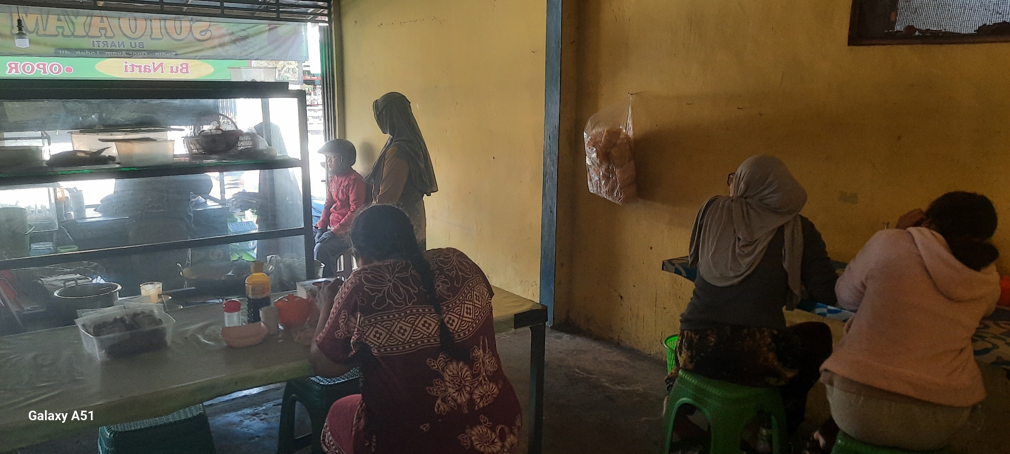 In Selopuro, Indonesia, a small food stall or eatery is depicted. The scene shows three women and a child. Two of the women are seated at a table on the right side of the image, facing away from the camera, with one wearing a light grey hijab and the other a pink hoodie. A third woman, wearing a dark maroon patterned dress and with her hair in a braid, is seated at a table on the left, also facing away. A young child in a red shirt and helmet is standing to her left, looking towards the food display. The eatery features a glass display case filled with food items, likely for sale. A yellow wall dominates the background, with a blue vertical beam separating sections of the establishment. A plastic bag filled with snacks hangs on the wall to the right. The lighting suggests an indoor setting, possibly during daylight hours.