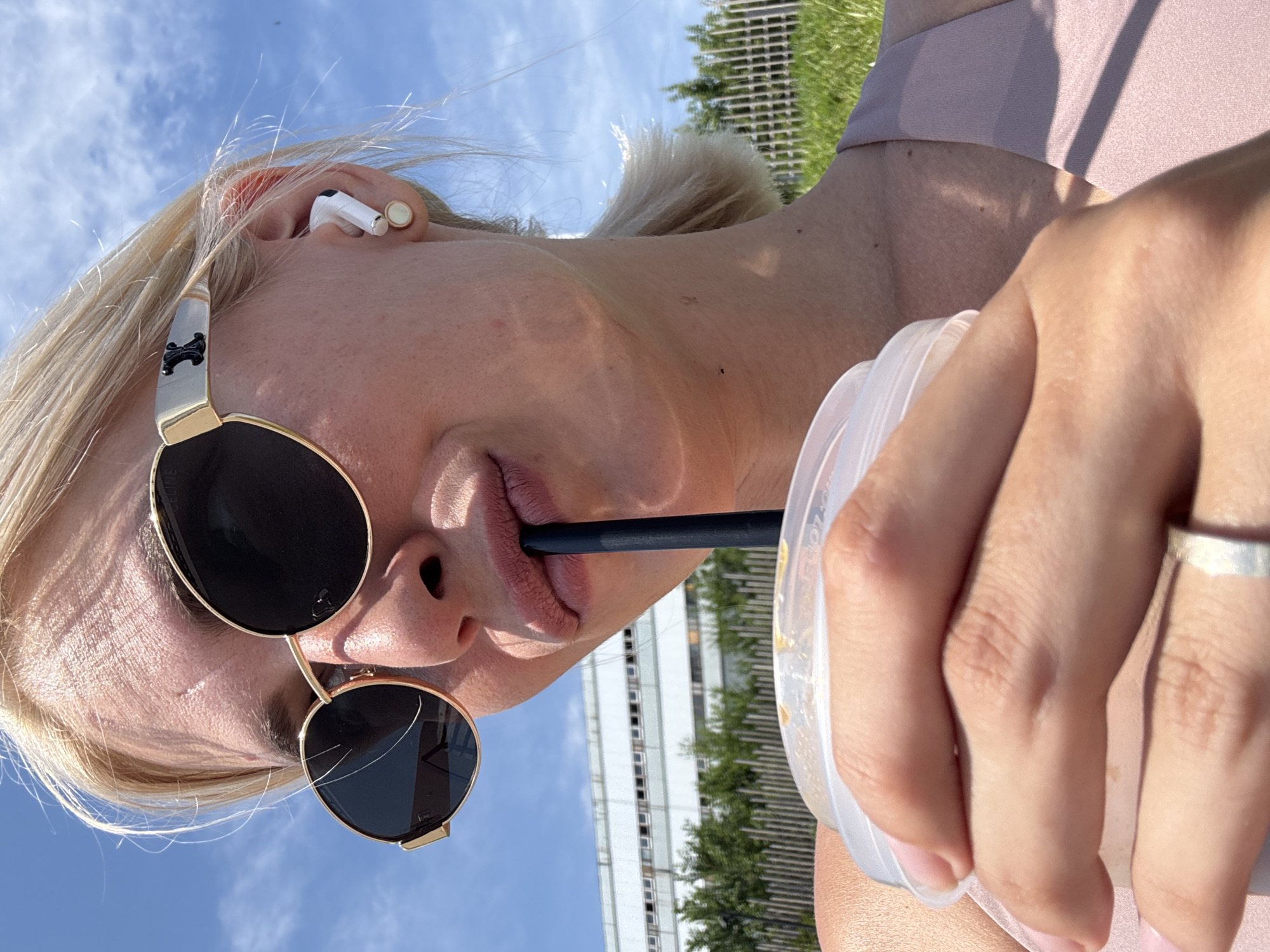 A young blonde woman is pictured outdoors, holding a clear plastic cup with a dark straw and drinking from it. She is wearing oversized, round sunglasses with gold frames and a black AirPods Pro in her right ear. The background consists of a blue sky with some clouds, a building with multiple windows, green trees, and a white picket fence. The woman is wearing a light pink top. The image is a close-up shot, focusing on her face and upper body. The scene is set on a sunny day.