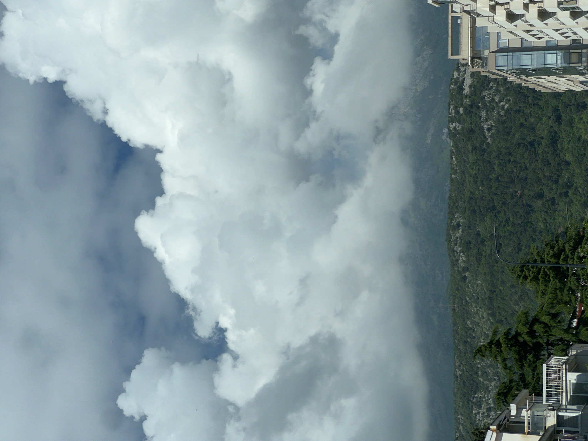 The image depicts a cloudy sky dominating the left side of the frame, contrasting with a green, densely vegetated mountain slope on the right. In the top right corner, a tall, modern building with multiple balconies is visible. Further down the mountain, a smaller building is partially obscured by trees. A utility pole curves across the lower portion of the mountain. The image appears to be taken from or near Budva, Montenegro, based on the terrain and building styles typically found there.