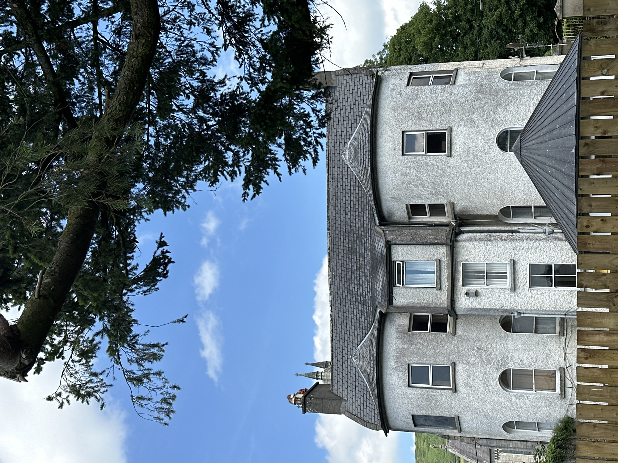 A large, two-story grey building with a grey slate roof and turrets sits in front of a blue sky with white clouds. A large tree branch with green needles hangs over the building from the top left corner of the image. The building has numerous windows, some with white frames and others with dark frames. A chimney with red brick trim is visible on the left side of the roof. In the background, a church steeple is visible. In the foreground, a wooden fence runs along the bottom of the image. The building is located in Baltinglass, Ireland.