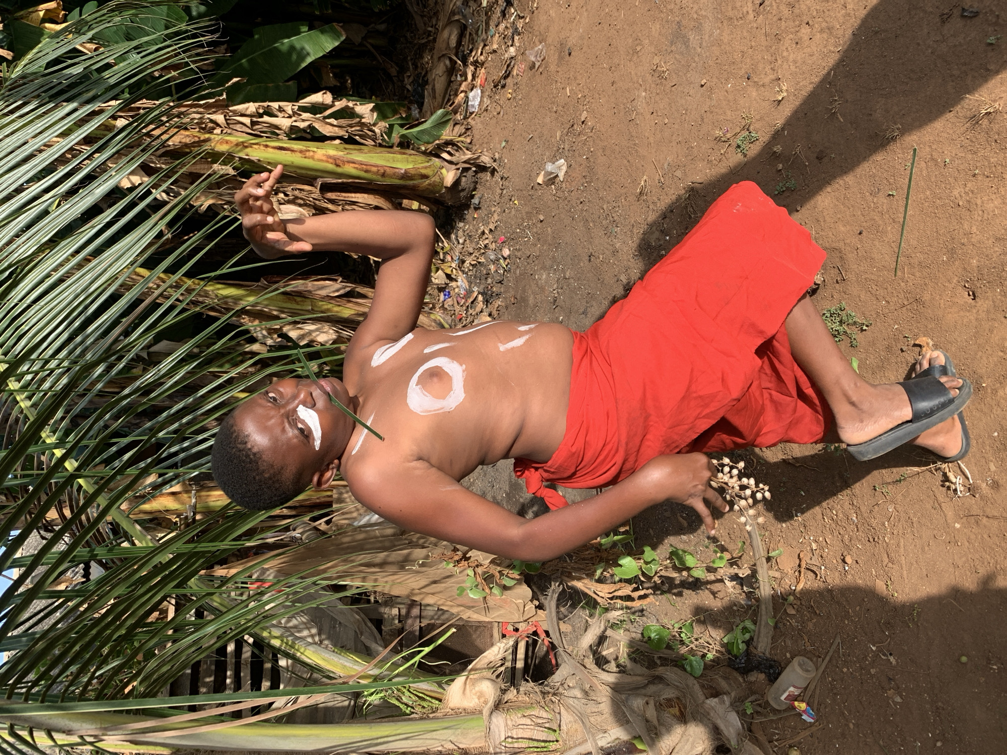 A shirtless individual, with white markings on the face and chest, lies on the ground. The person is wearing a red cloth wrapped around the lower body and black sandals on the feet. The setting appears to be outdoors, with palm leaves, banana plant debris, and dry ground visible. A thin, green stalk rests on the person's chest. The ground has scattered debris, including plastic and dried vegetation. The image was taken in Ikorodu, Nigeria.